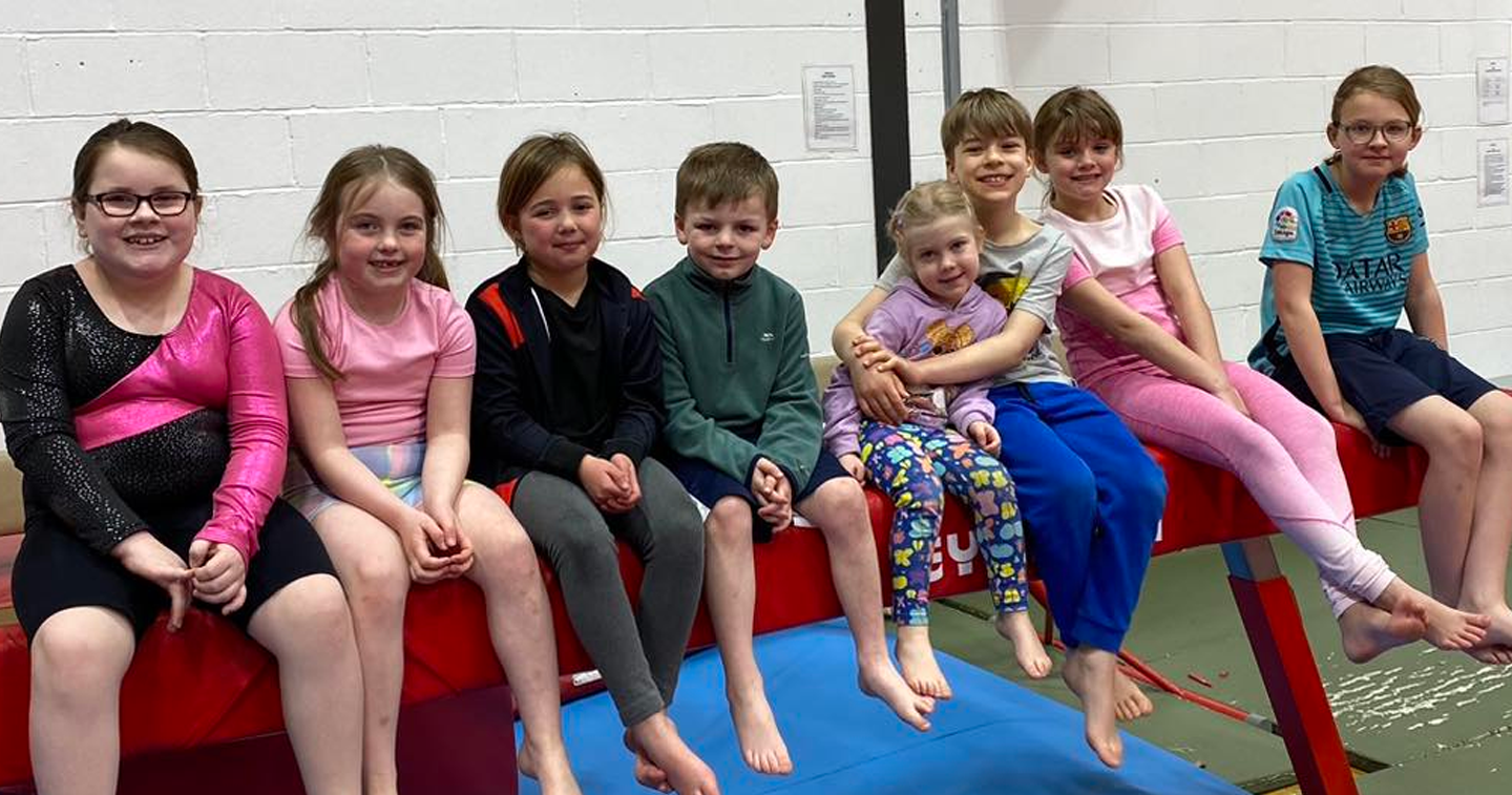 A group of children are sitting on a balance beam in a gym.
