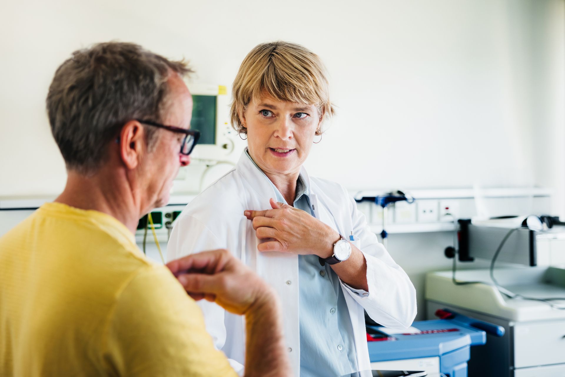 A female mature doctor, in a white coat, speaks with a patient in a hospital room.