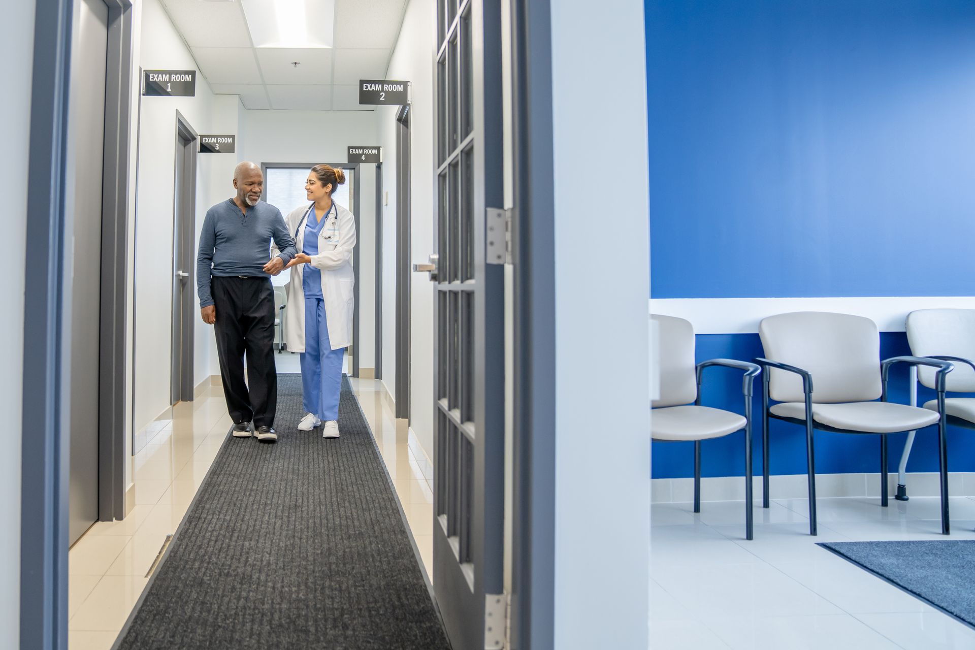 Doctor helping patient in clinic hallway near exam rooms and waiting area.