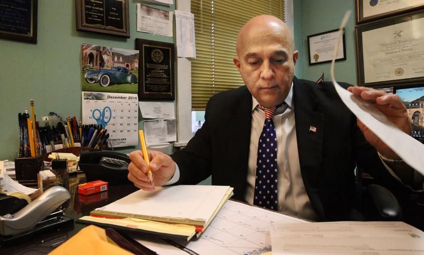 A man in a suit and patterned tie sits at a desk, writing in a notebook while holding papers in a busy office setting