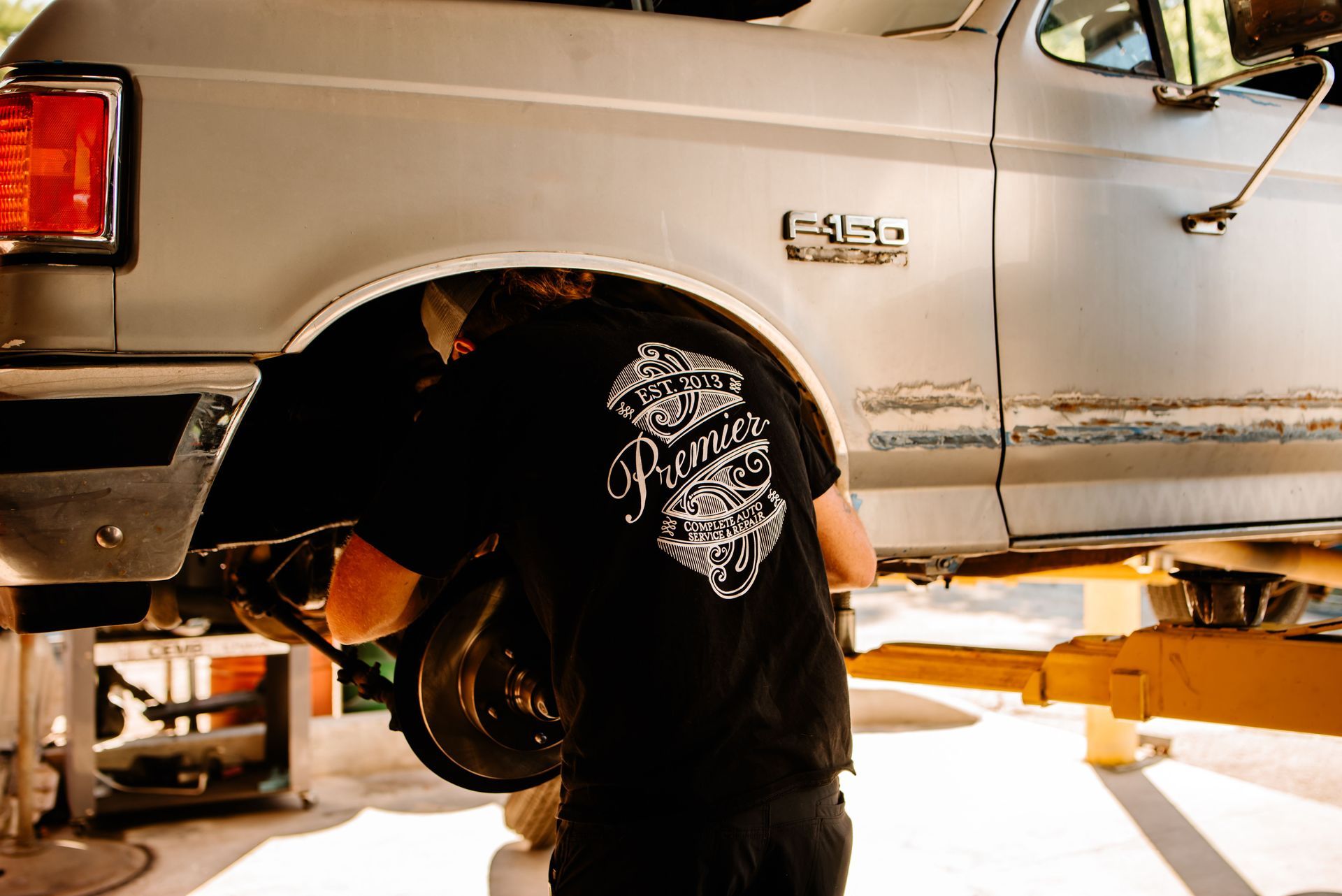 Man with Premier Auto Care work shirt on, working under vehicle on the brakes of an old truck.