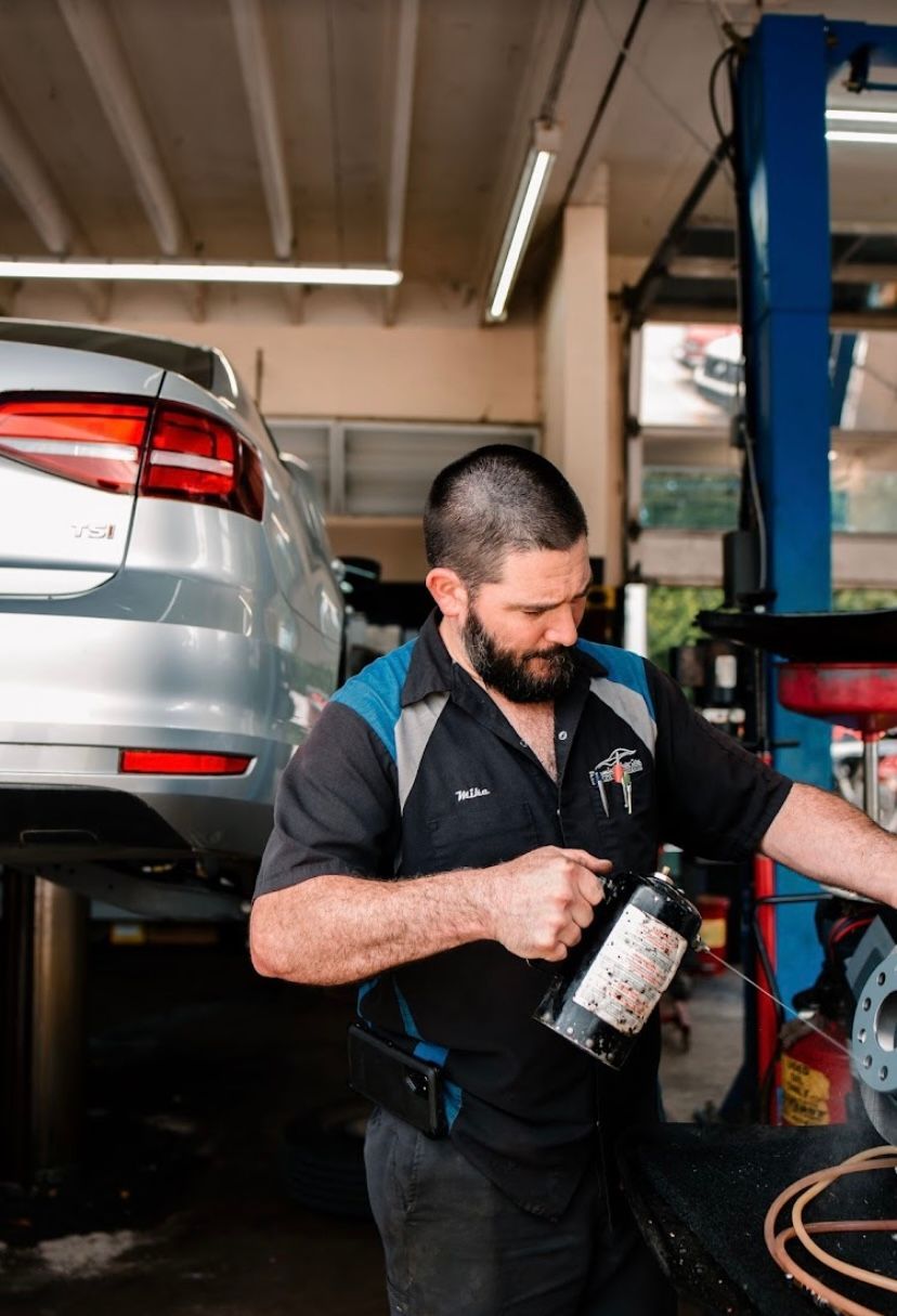 A man is working on a car in a garage while holding a bottle of wine.