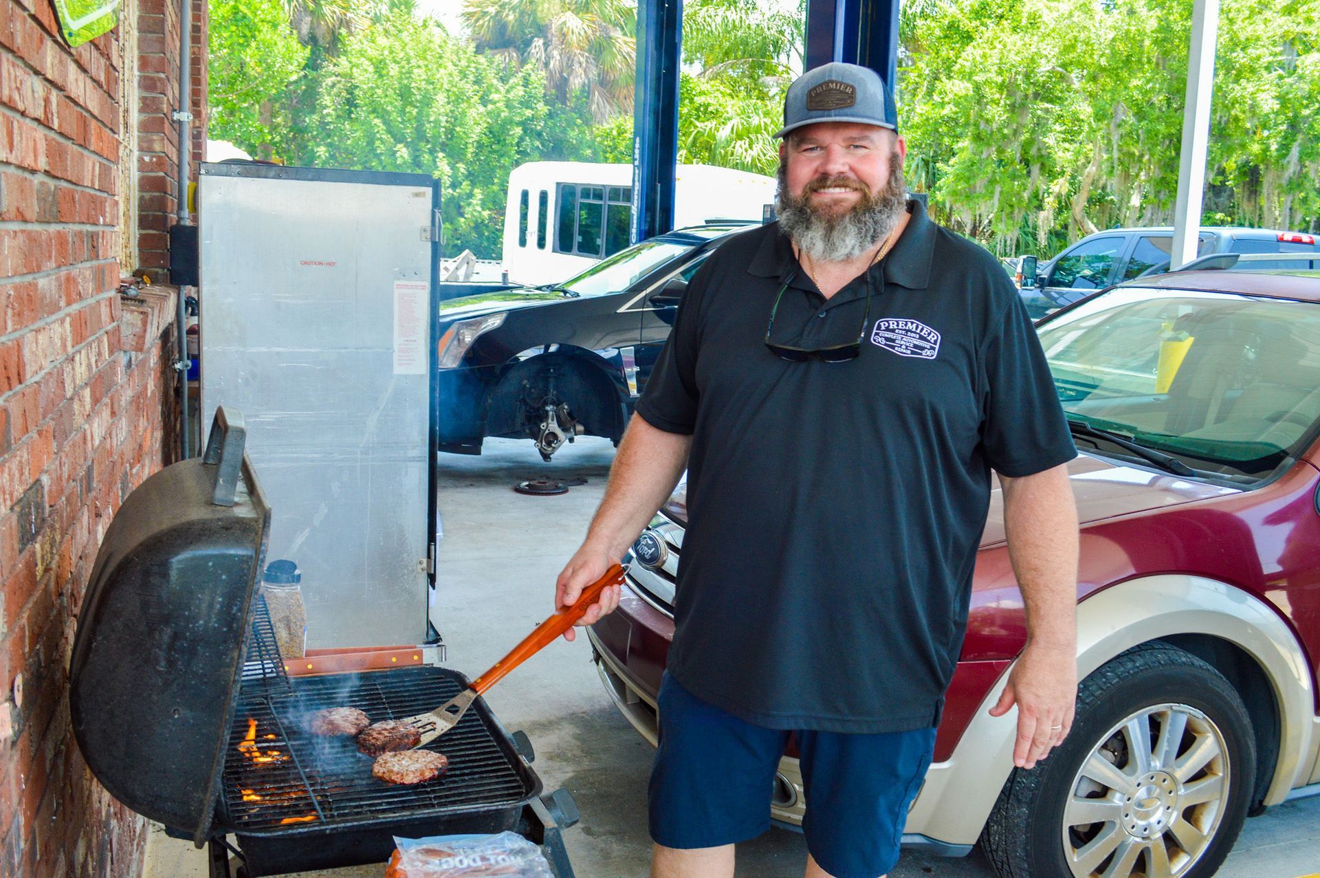 A man with a beard is grilling hamburgers on a grill in front of a car.