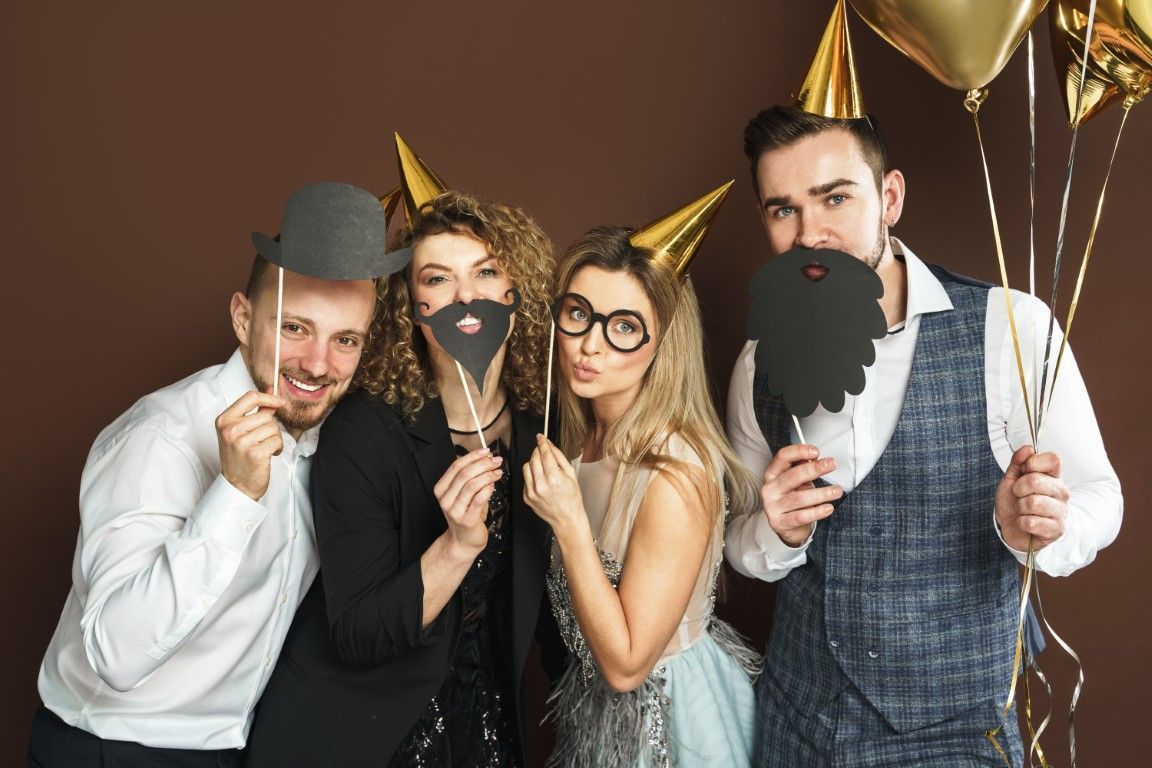 Four people in party hats posing with fake beards and glasses, holding gold balloons against a brown backdrop.
