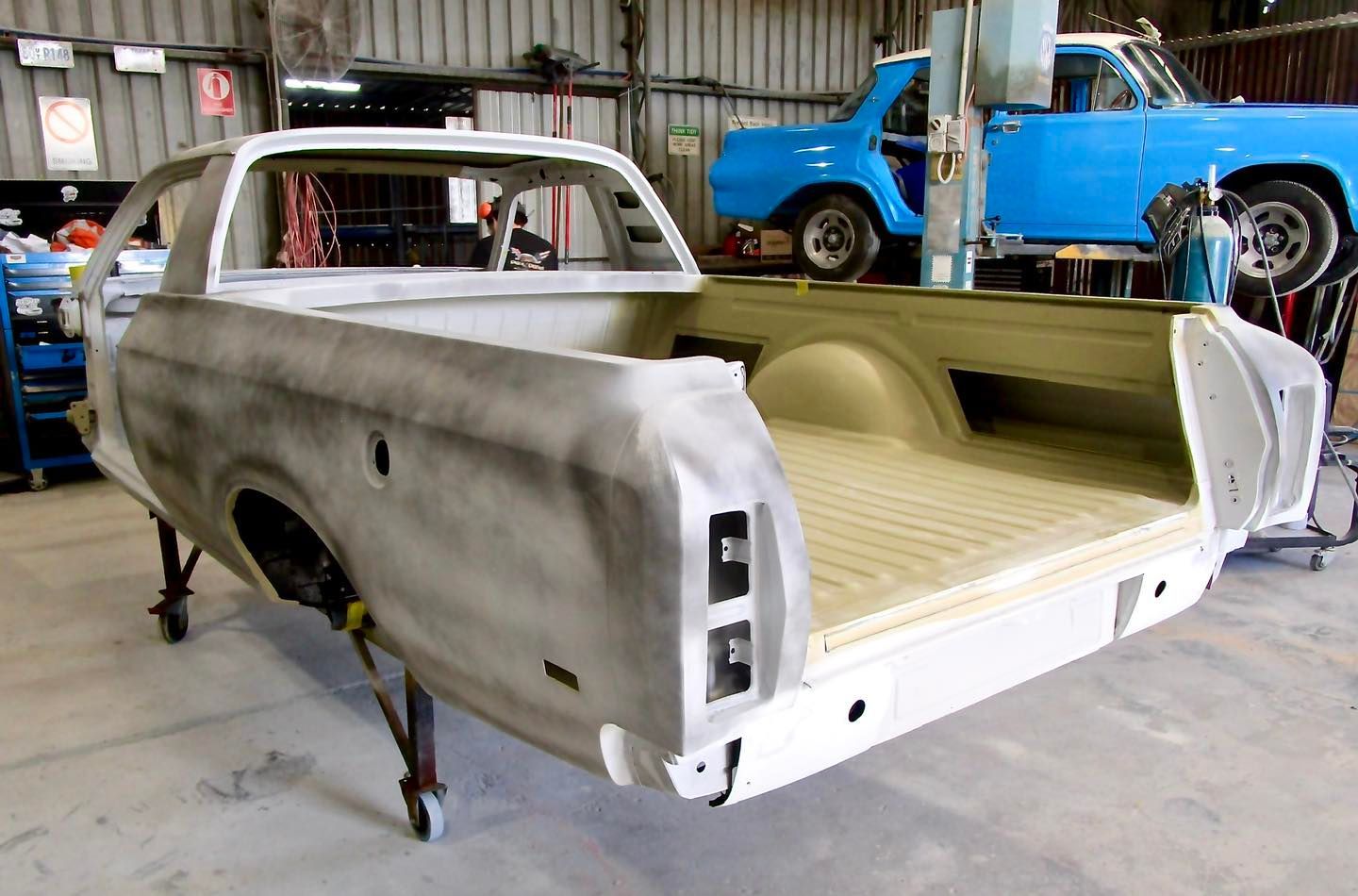 Partially restored white pickup truck in a repair shop; blue car on lift in background.