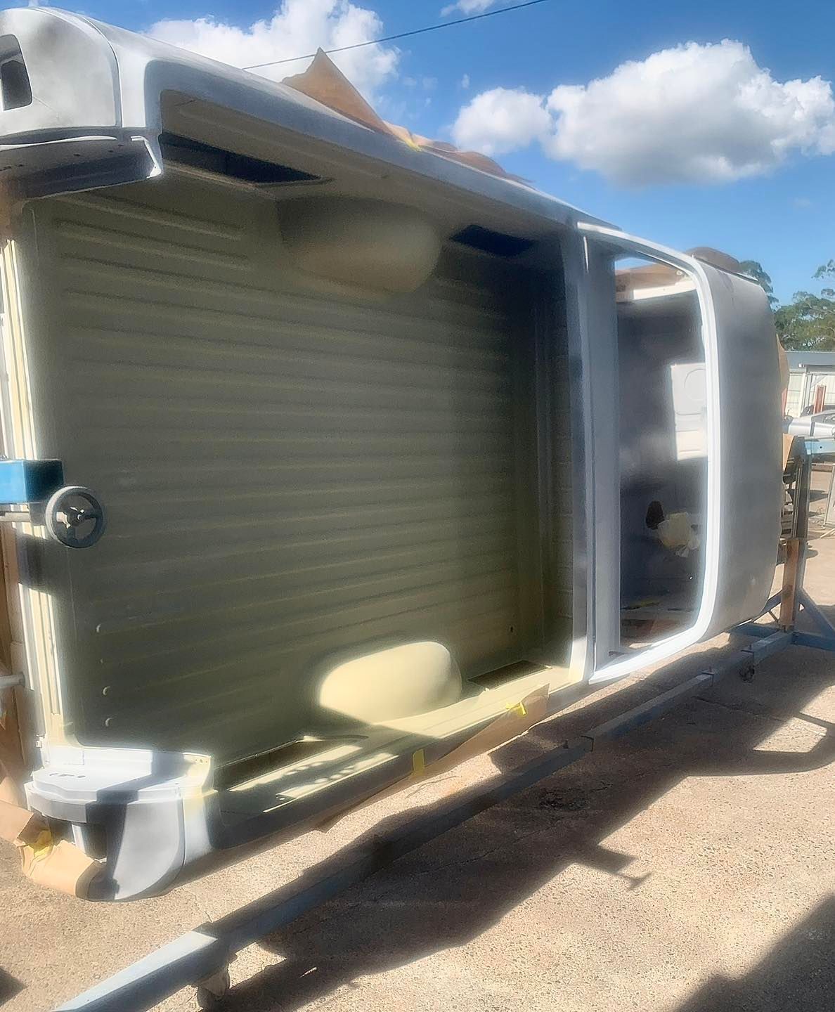 Upside-down car body on a rotisserie. Primarily gray and tan in a sunny outdoor setting.