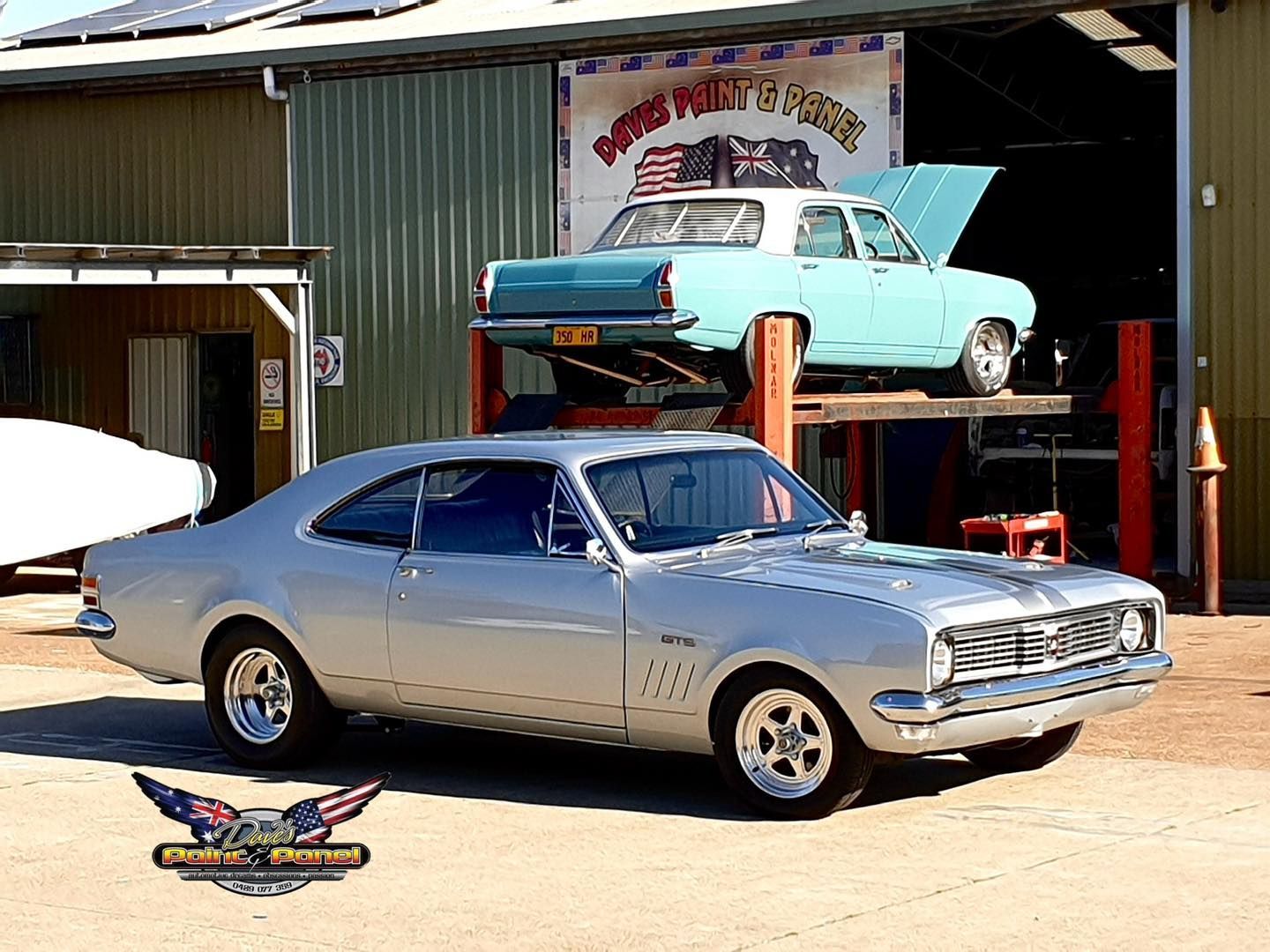Silver muscle car parked in front of a garage, blue classic car lifted above.