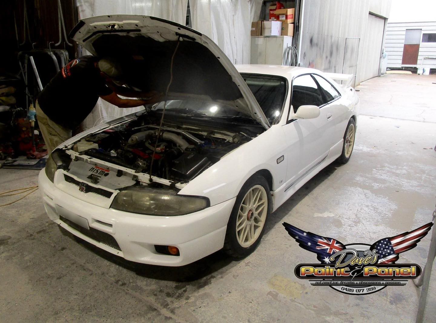 White Nissan Skyline with the hood open in a garage setting; a person works on the engine.