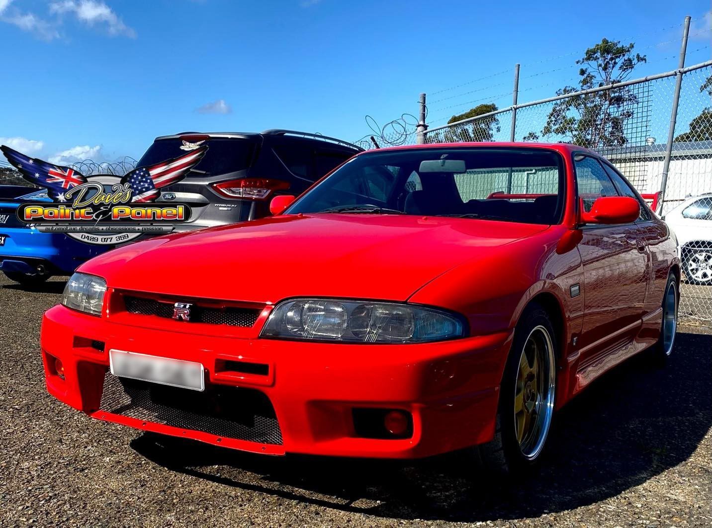 Red Nissan Skyline R33 sports car at an outdoor event, with gold wheels and a bright blue sky.