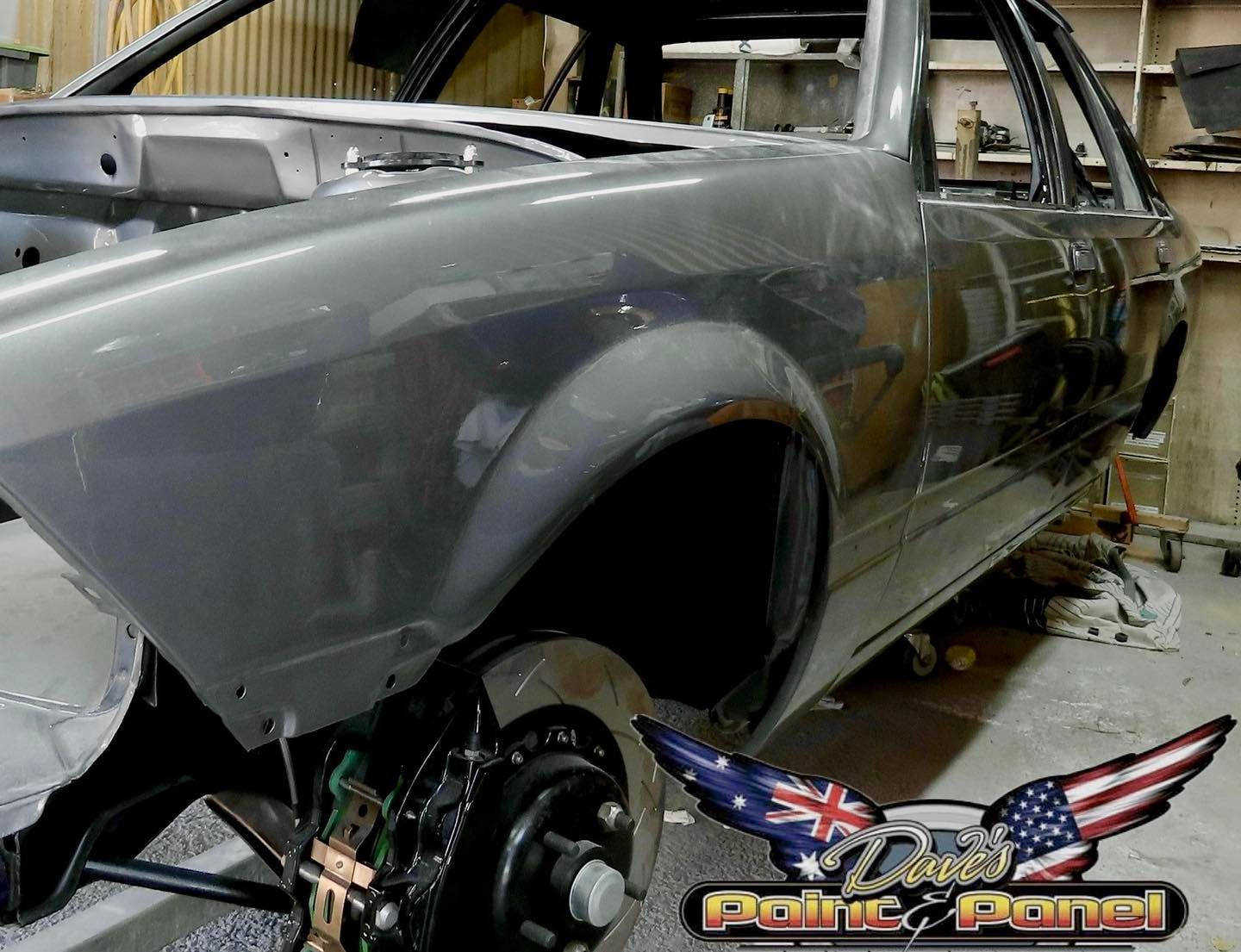 Gray car body in a workshop, front wheel visible, primed for paint.