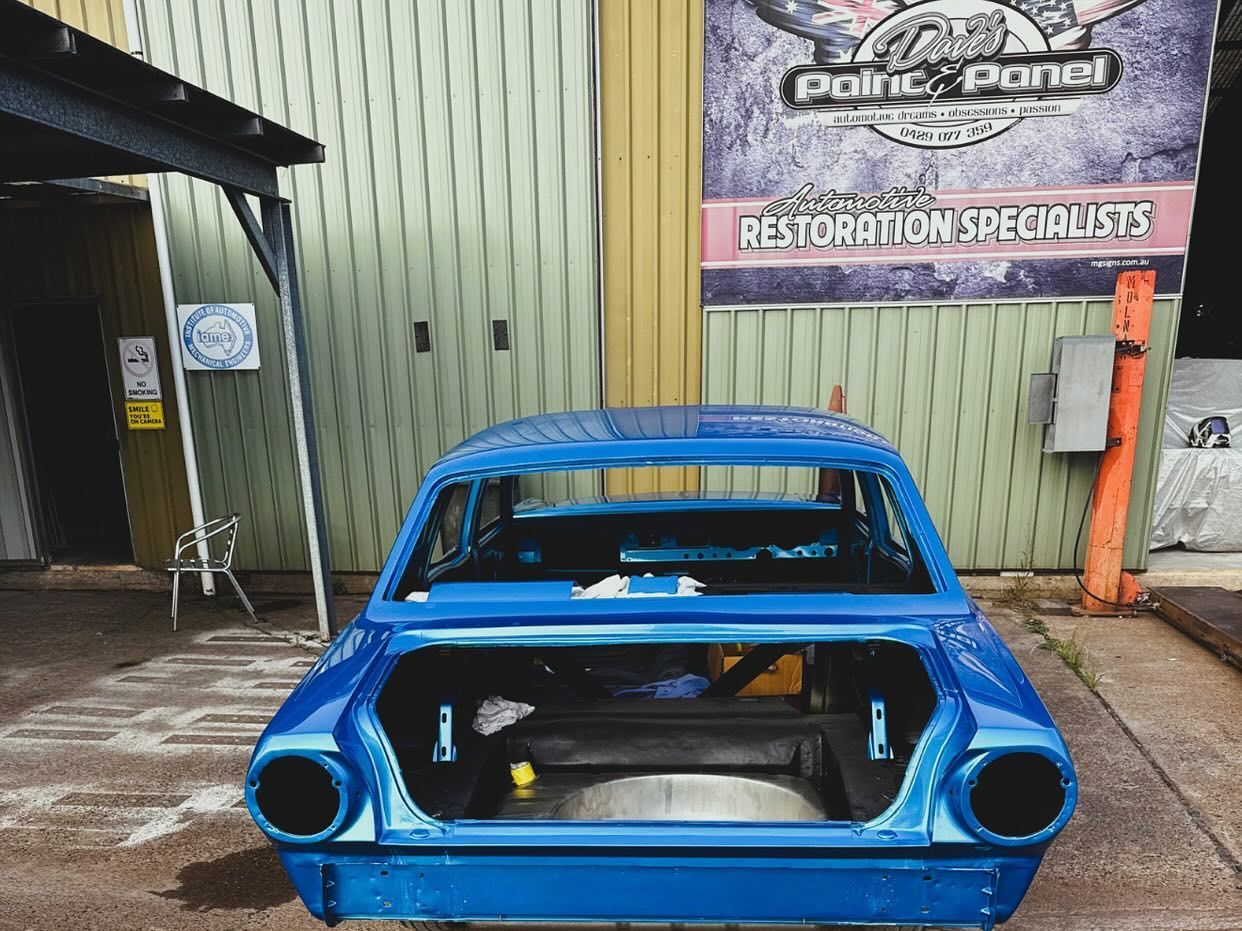 Blue car chassis in a shop bay, showing rear. Restoration shop sign in the background.