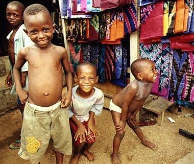 Quattro bambini sorridenti davanti a un negozio di tessuti colorati. Uno di loro ha il torso nudo.