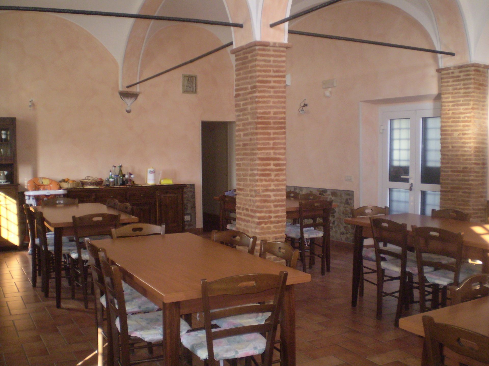 Sala da pranzo con tavoli in legno, sedie e colonne in mattoni. Illuminazione calda.