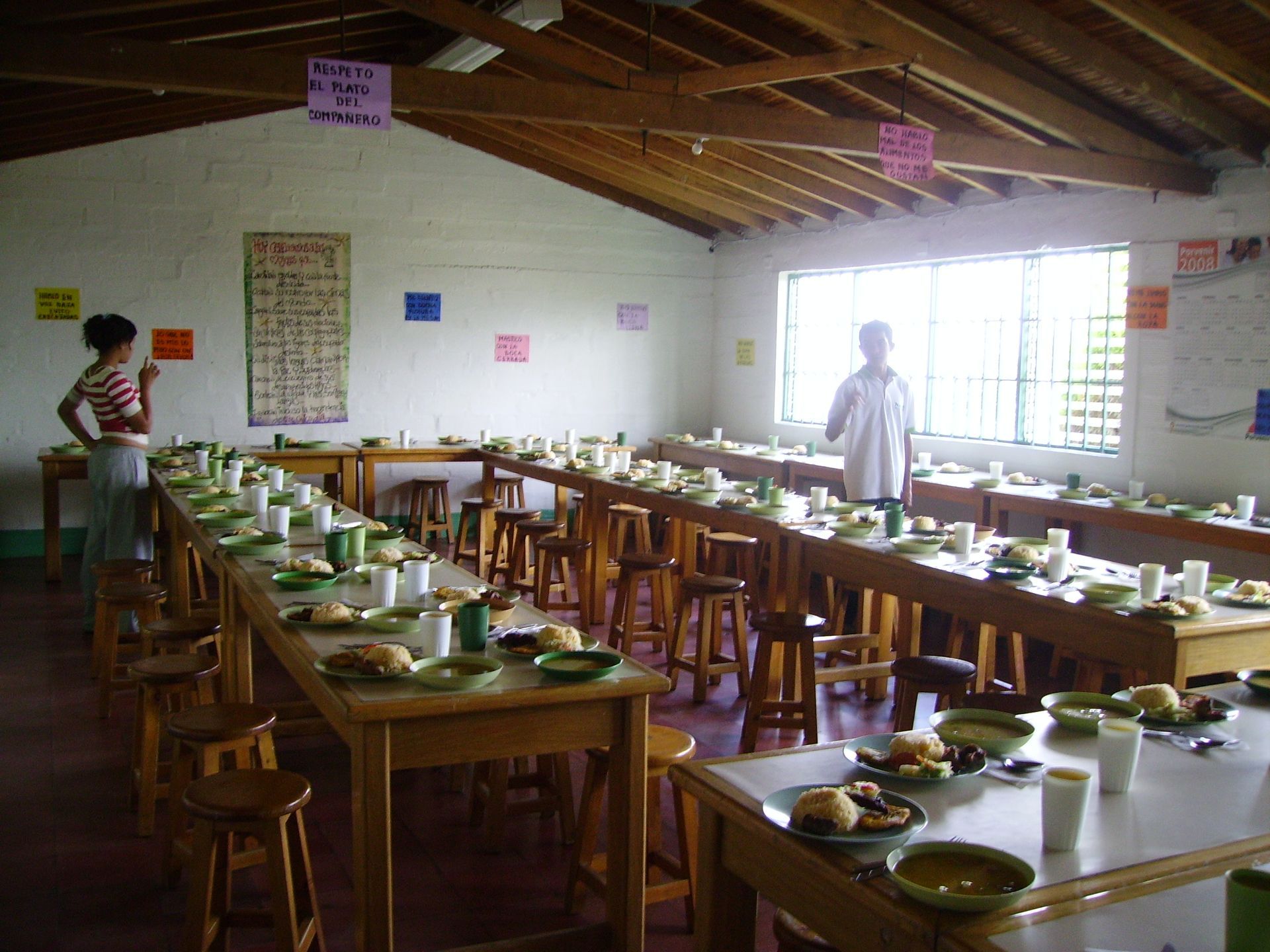Lunghi tavoli apparecchiati per un pasto in una semplice sala da pranzo. Due persone sono in piedi vicino ai tavoli, in attesa dei commensali.