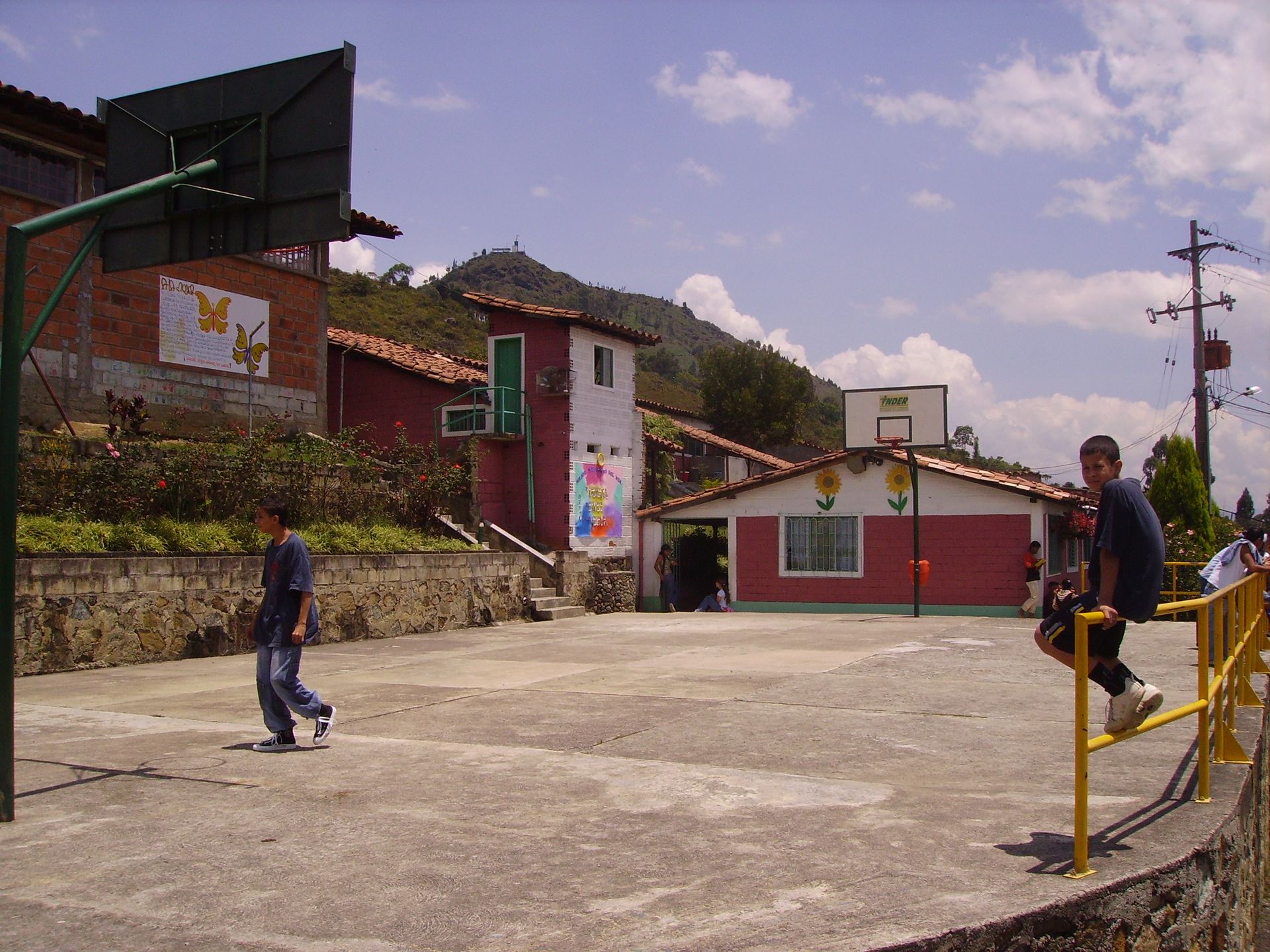 Due ragazzi su un campo da basket. Edifici con murales, montagne sullo sfondo. Giornata di sole.