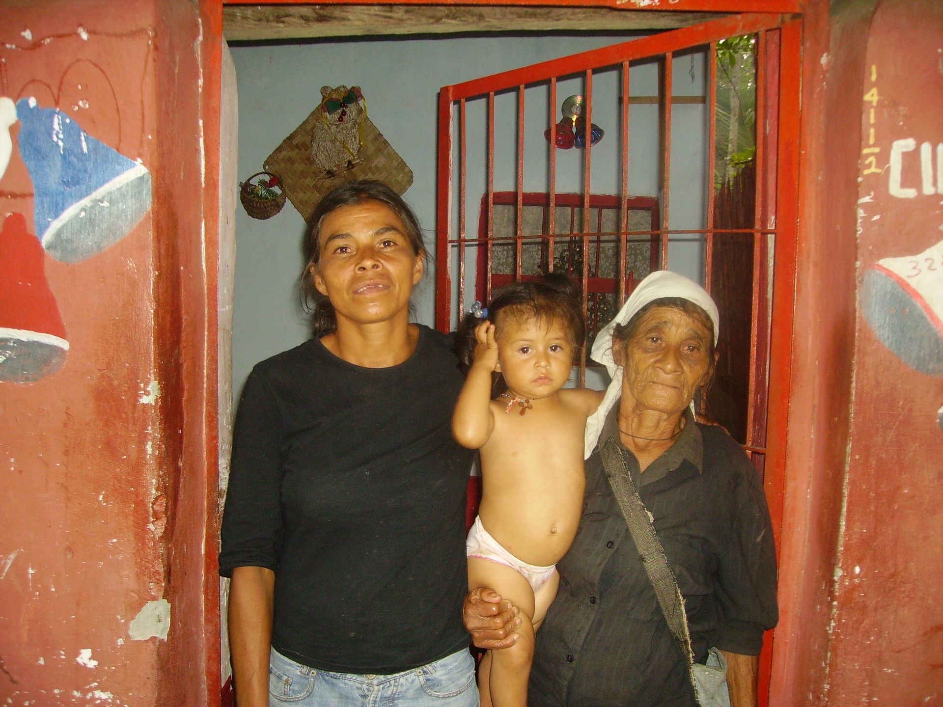 Tre persone posano sulla soglia: una donna, un bambino e una donna anziana. Lo stipite della porta è rosso, le pareti sono dipinte.