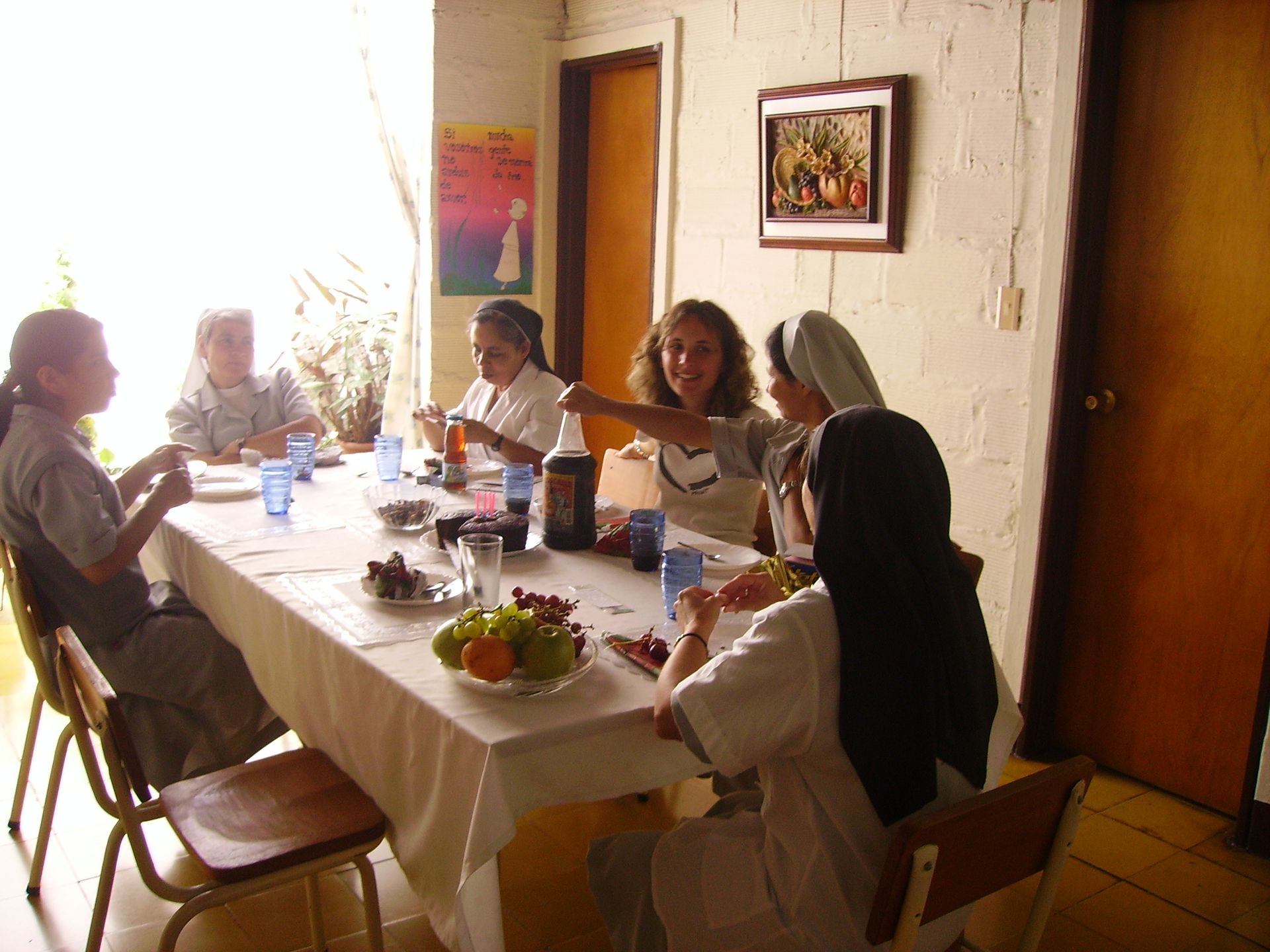 Persone sedute a un tavolo che mangiano, una stanza luminosa. Una donna che indica, altri individui che sorridono.