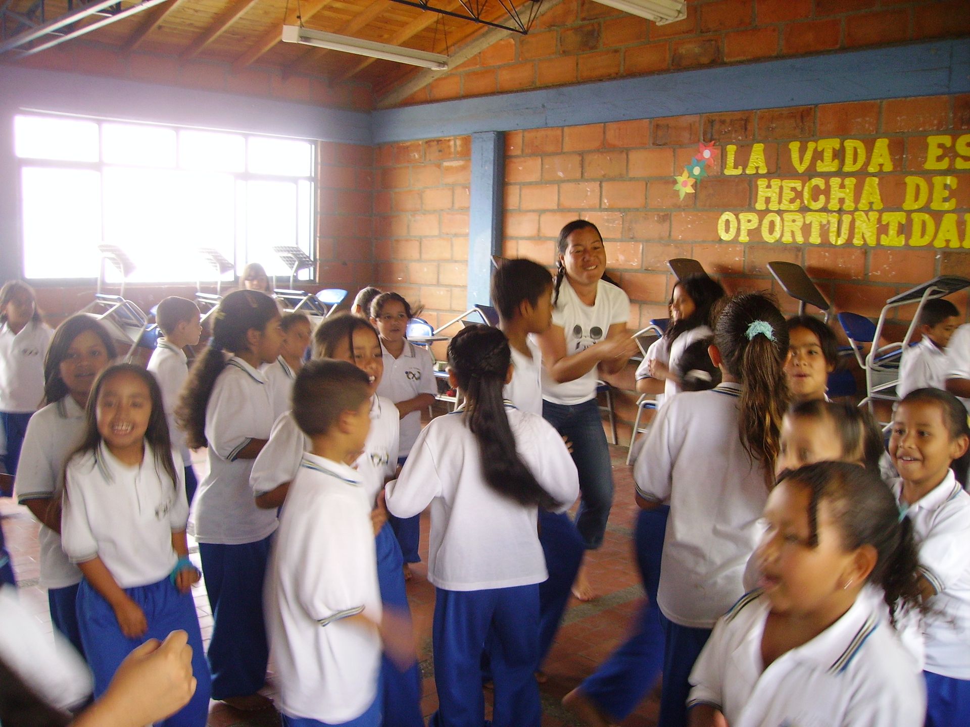 Bambini in uniforme blu e bianca giocano in una stanza, guidati da una donna. Pareti di mattoni, testo in spagnolo.
