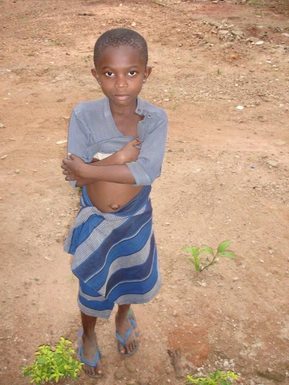 Un ragazzo è in piedi all'aperto con le braccia incrociate, indossa una sciarpa blu e una maglietta grigia. È circondato da terra marrone.