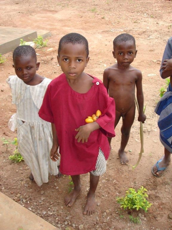 Bambini in piedi all'aperto, sulla terra; uno indossa il rosso, un altro il bianco e un terzo è a torso nudo.