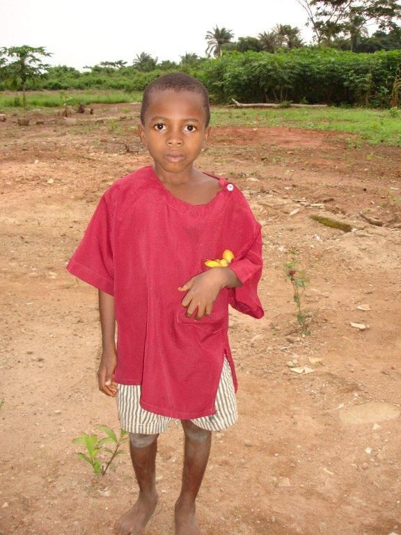 Un giovane è in piedi in un campo sterrato, indossa una maglietta rossa e pantaloncini a righe. È a piedi nudi.