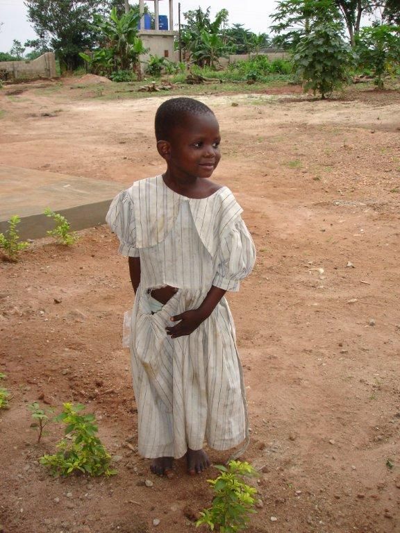 Ragazza che indossa un vestito chiaro, in piedi all'aperto su un terreno sterrato, che guarda di lato con un leggero sorriso.