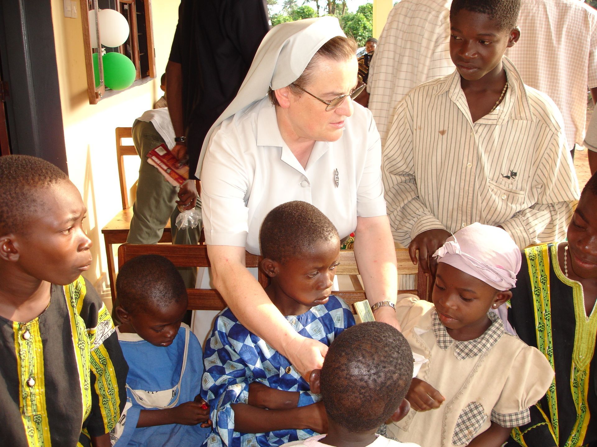 Una suora circondata da bambini interagisce all'aperto. Alcuni bambini sono seduti, altri in piedi.