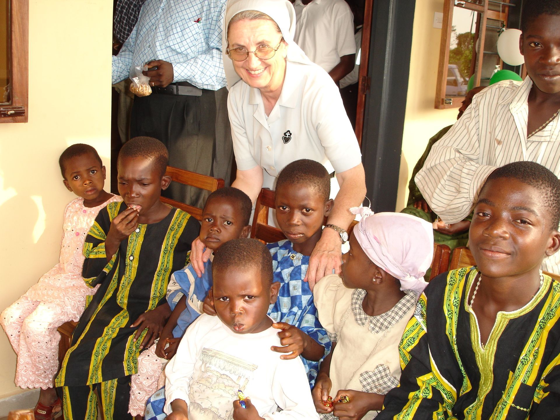 Una suora è circondata da diversi bambini. Sono seduti e sorridono.