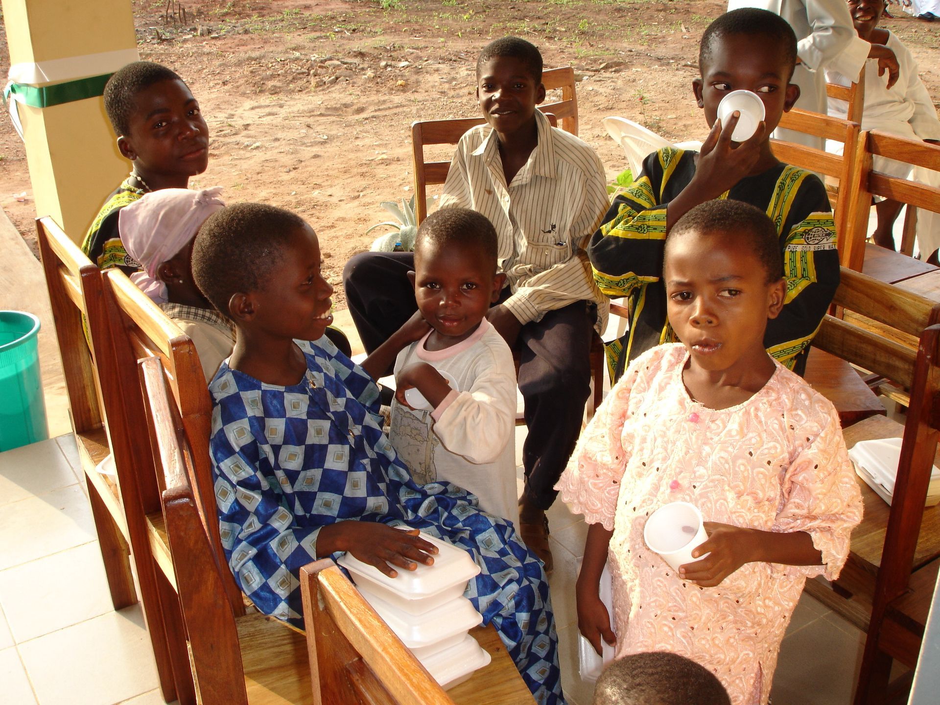 Bambini in un ambiente clinico. Alcuni tengono in mano bicchieri di carta, altri sono seduti su sedie di legno.