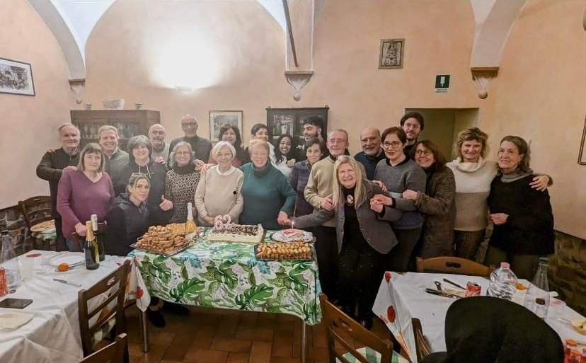 Un gruppo di persone festeggia in un locale, in posa attorno a una torta di compleanno su un tavolo.