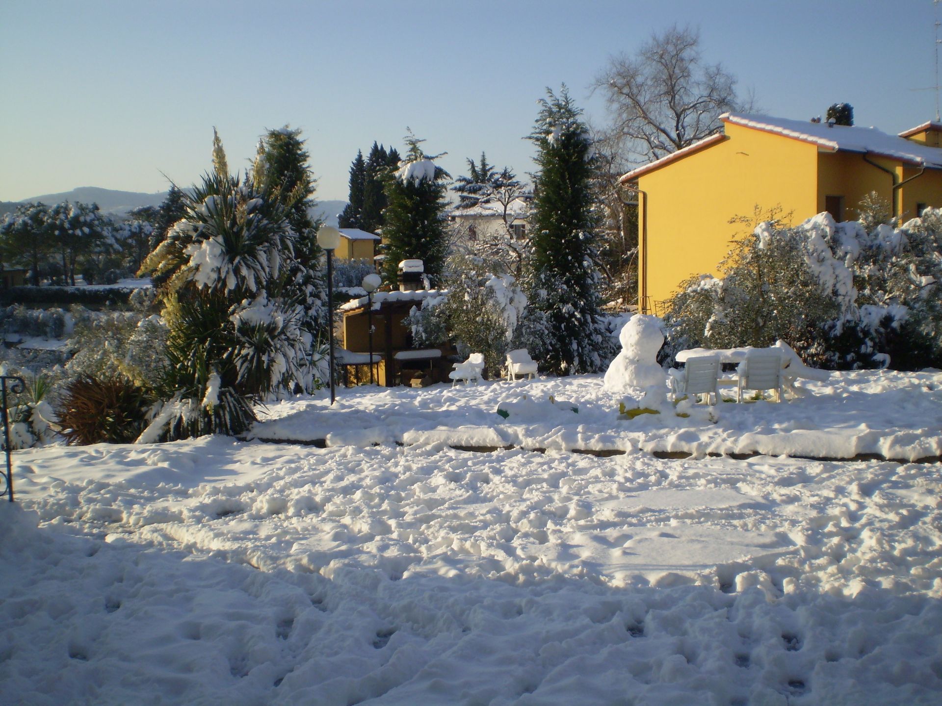 Cortile innevato con un pupazzo di neve davanti a un edificio giallo e alberi.