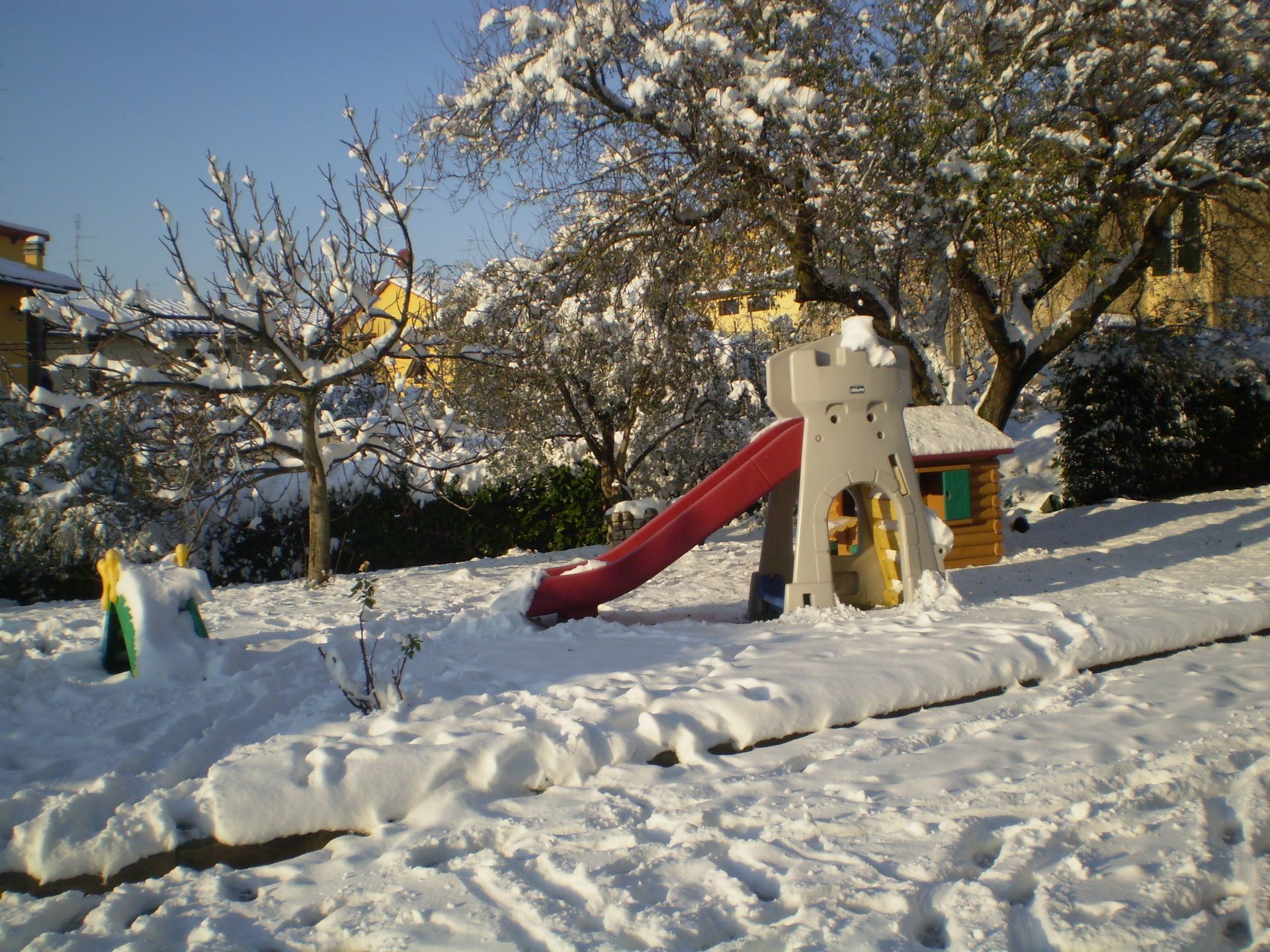 Parco giochi coperto di neve; scivolo, struttura a castello, piccola casa di legno, alberi e cielo azzurro.