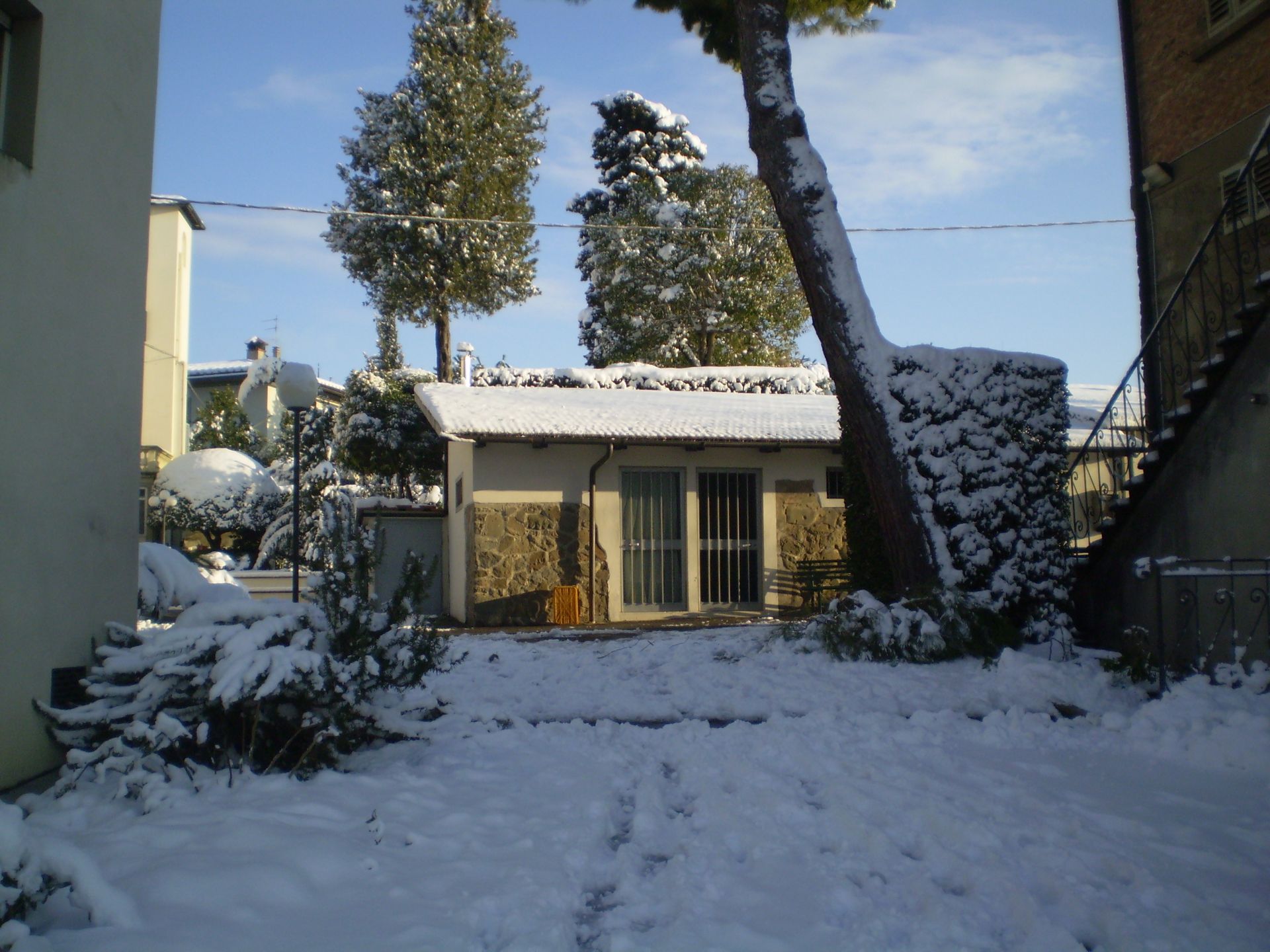 Cortile innevato con un piccolo edificio, alberi e impronte nella neve sotto un cielo luminoso.
