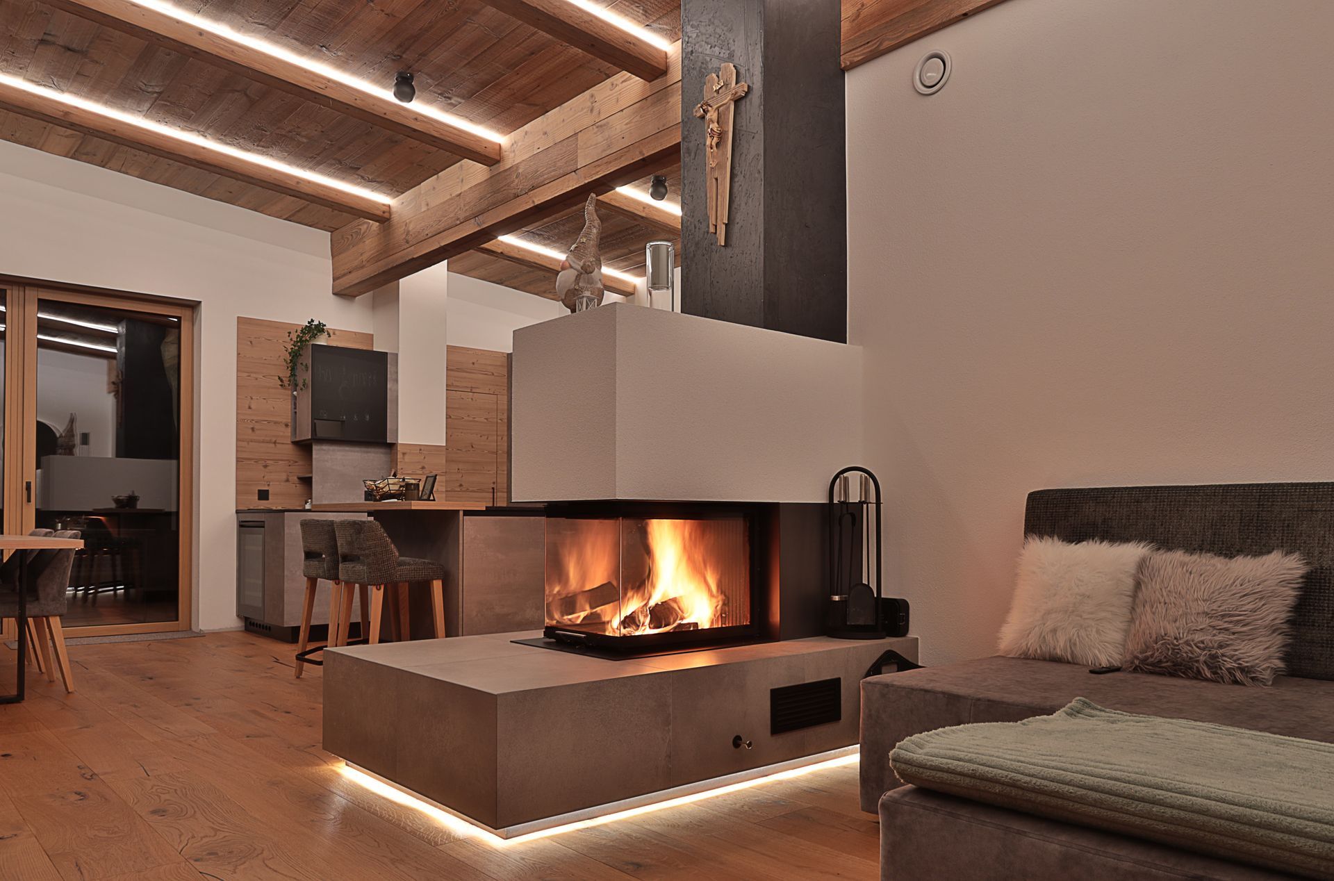 Gemütliches, modernes Wohnzimmer mit beleuchtetem Kamin, Holzbalken an der Decke und einem Sofa mit Fellüberwurf.