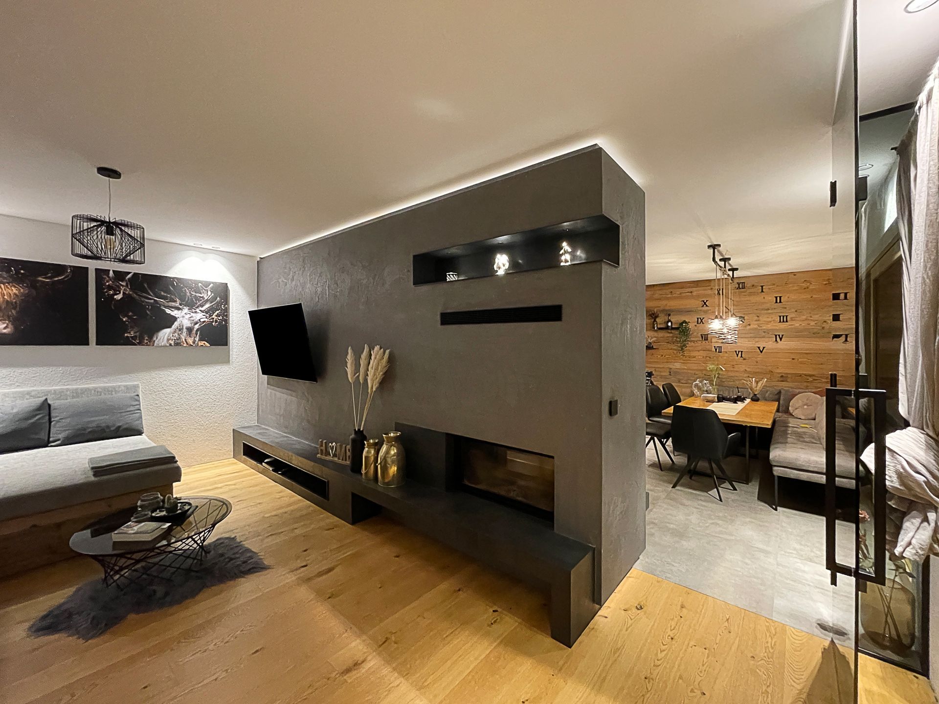 Modernes Wohnzimmer mit grauer Kaminwand, wandmontiertem Fernseher, Holzboden und Essbereich hinter einer Glastrennwand