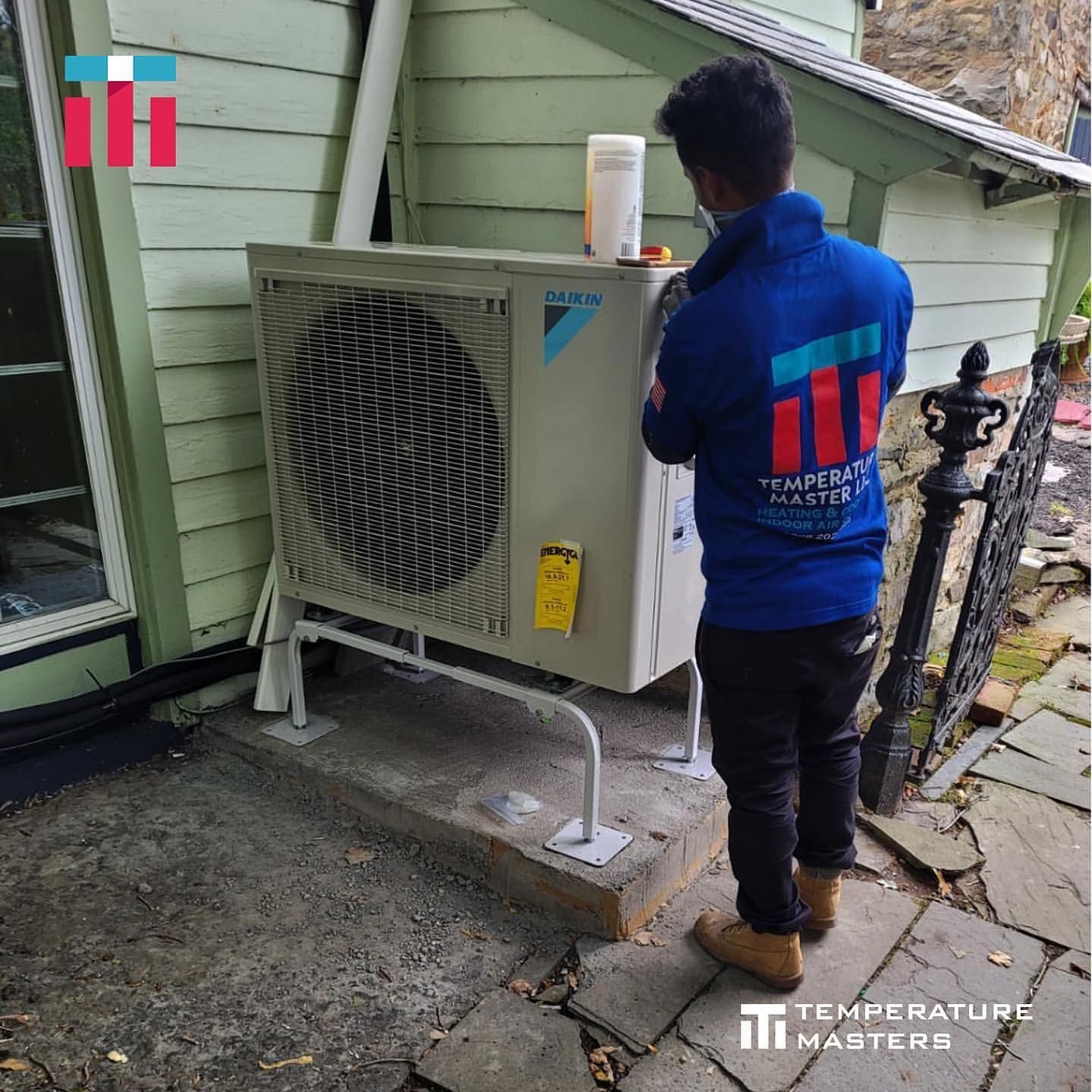 A man in a blue shirt is working on an air conditioner outside of a house.