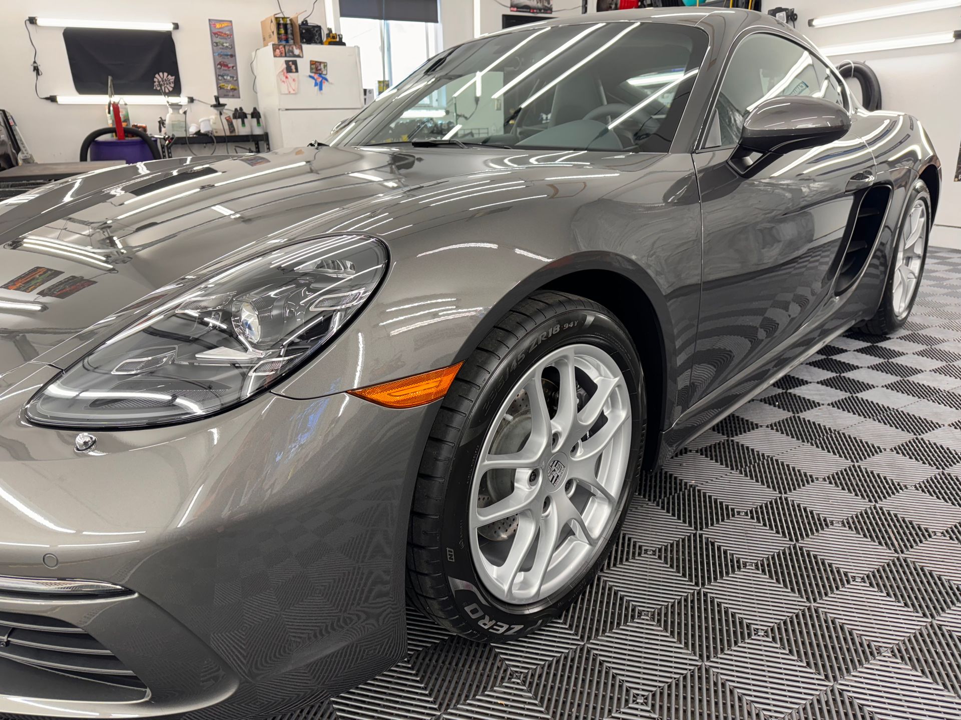 A gray porsche boxster is parked on a checkered floor in a garage.