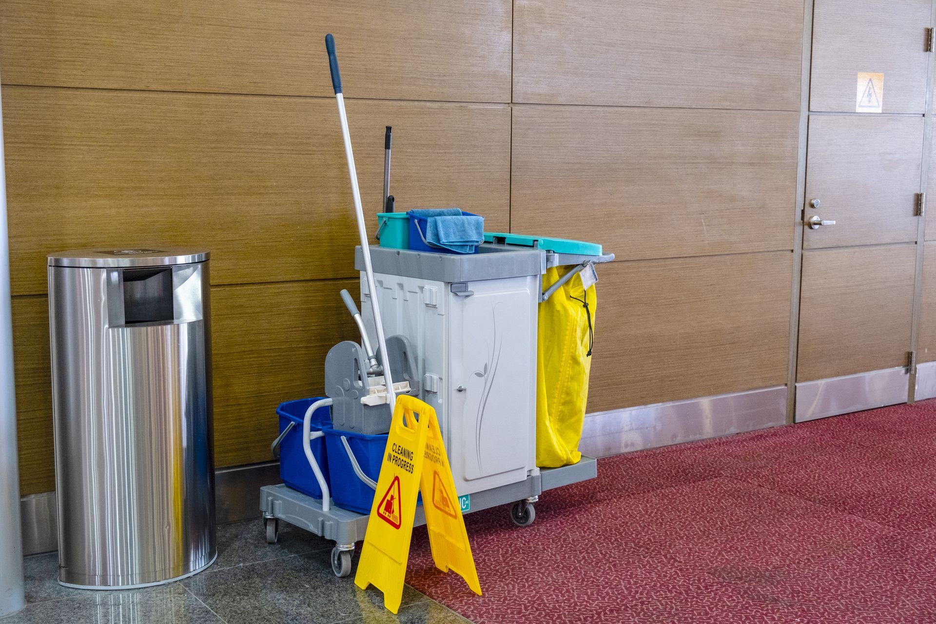 Cleaning cart with tools, cleaning buckets and equipment standing near commercial restroom door.
