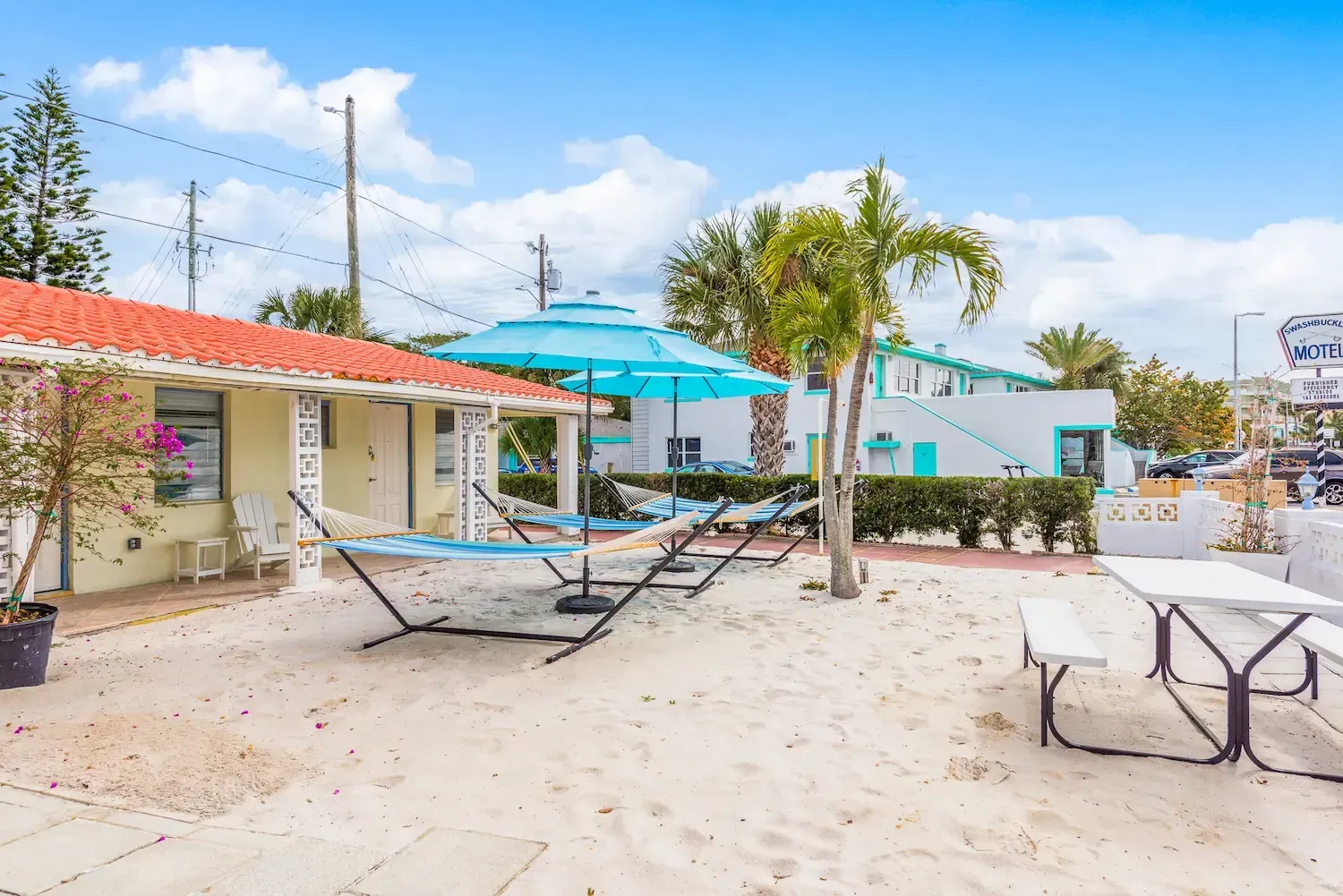 A picnic table and hammock are in the sand in front of a house.