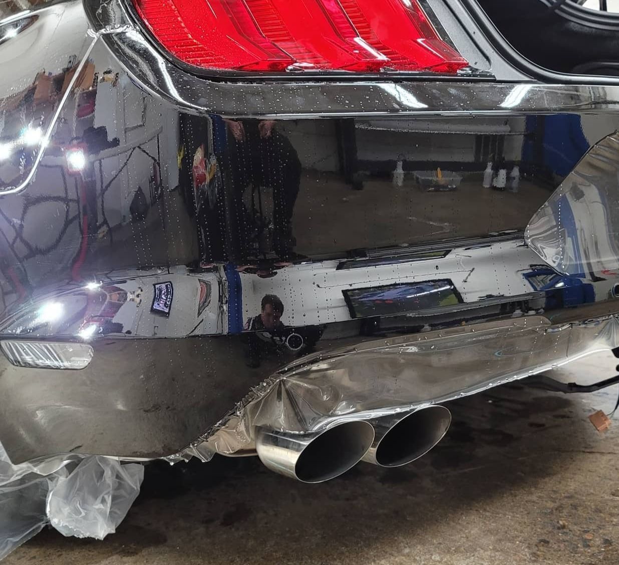 Close-up of a black car’s rear bumper with dual exhaust pipes and red taillight, showing damage and scuffs.