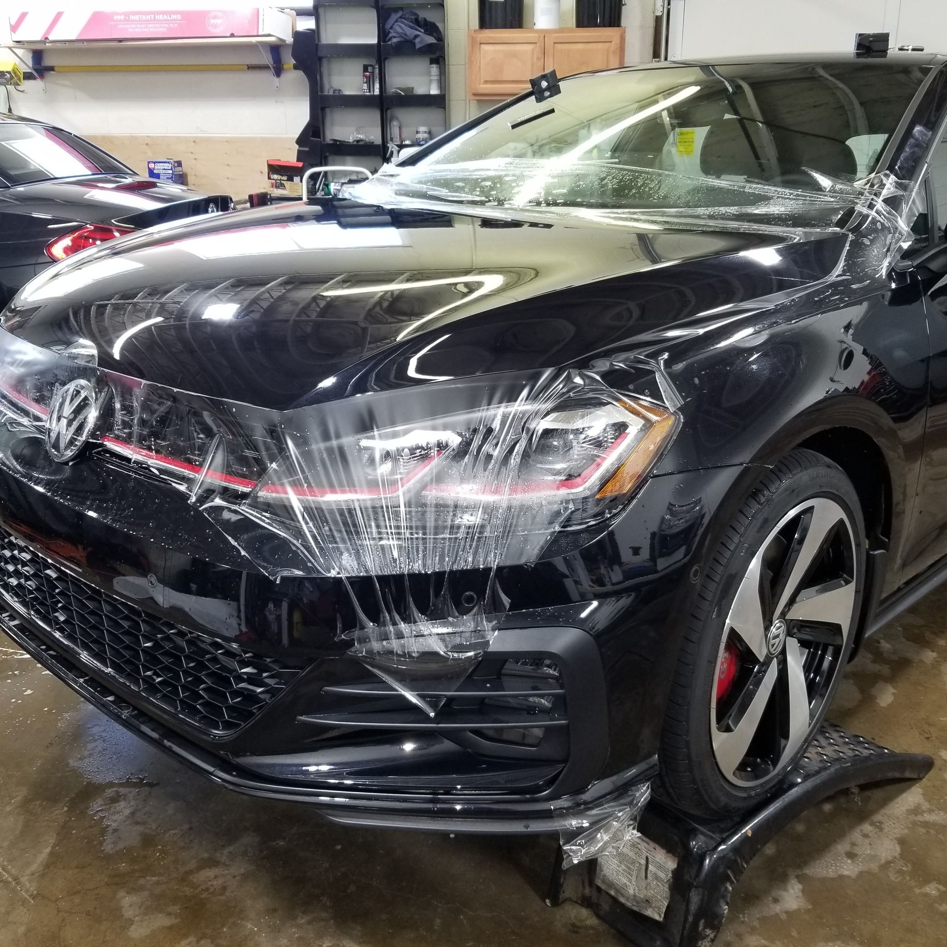 Black Volkswagen GTI with protective film being applied in a garage.