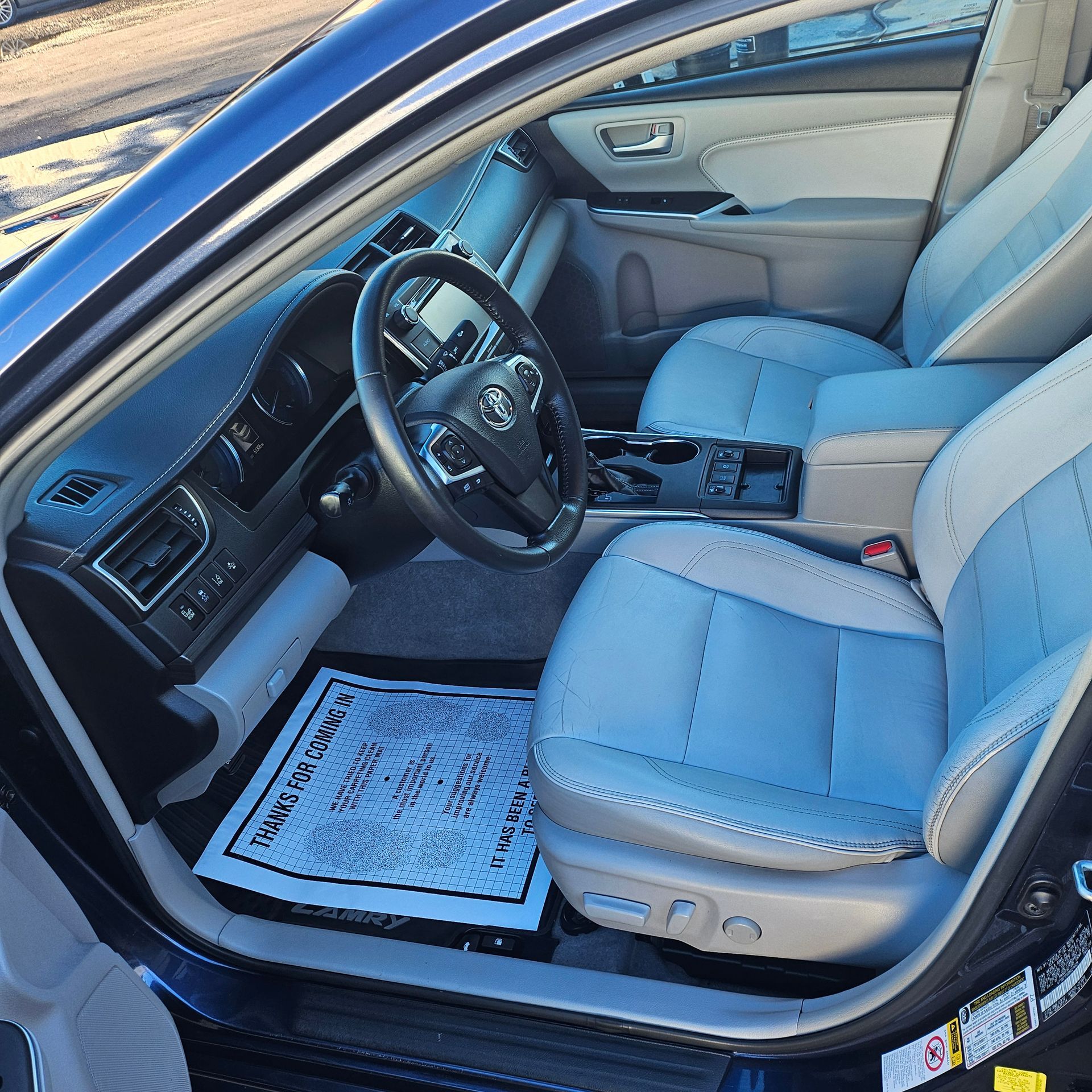 Blue car interior with beige leather seats, steering wheel, center console, and floor mat visible from the open driver’s door