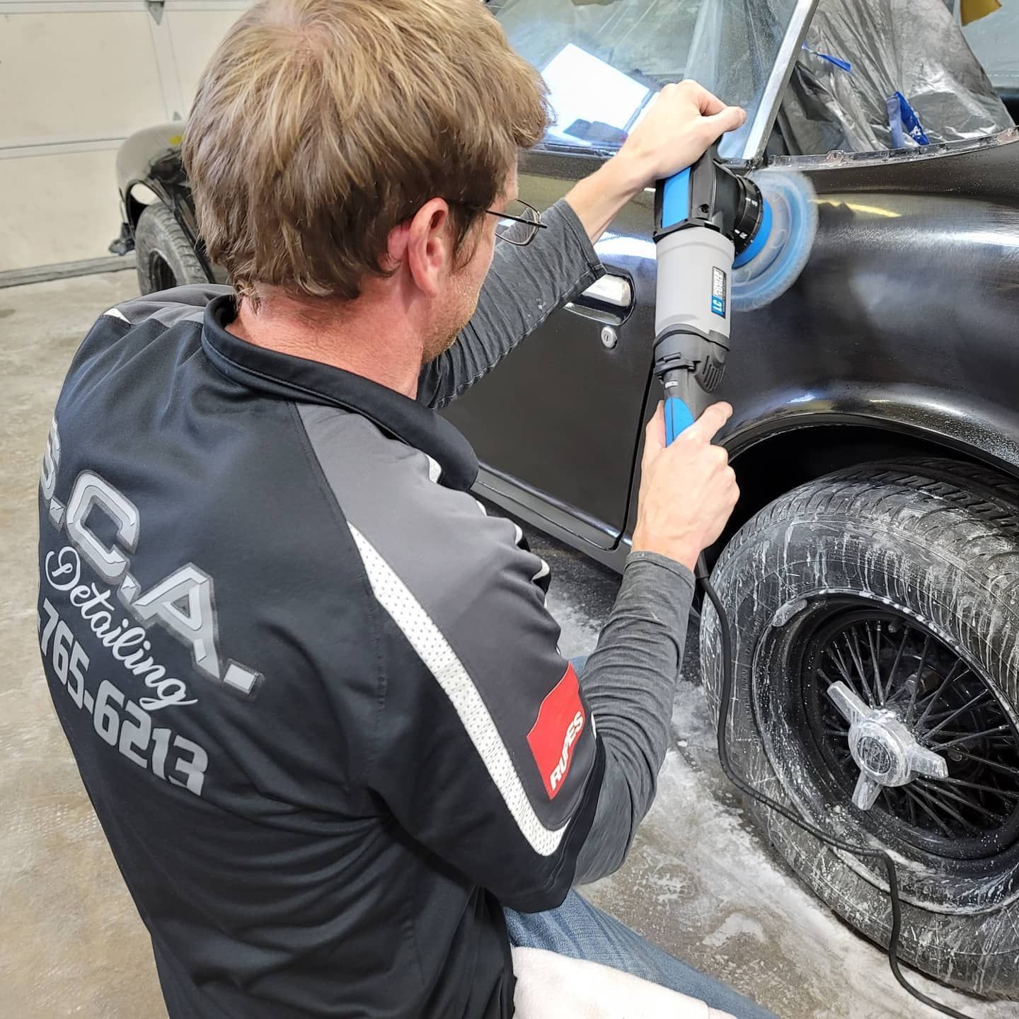 Man polishing a black car with a power tool, wearing a work shirt.