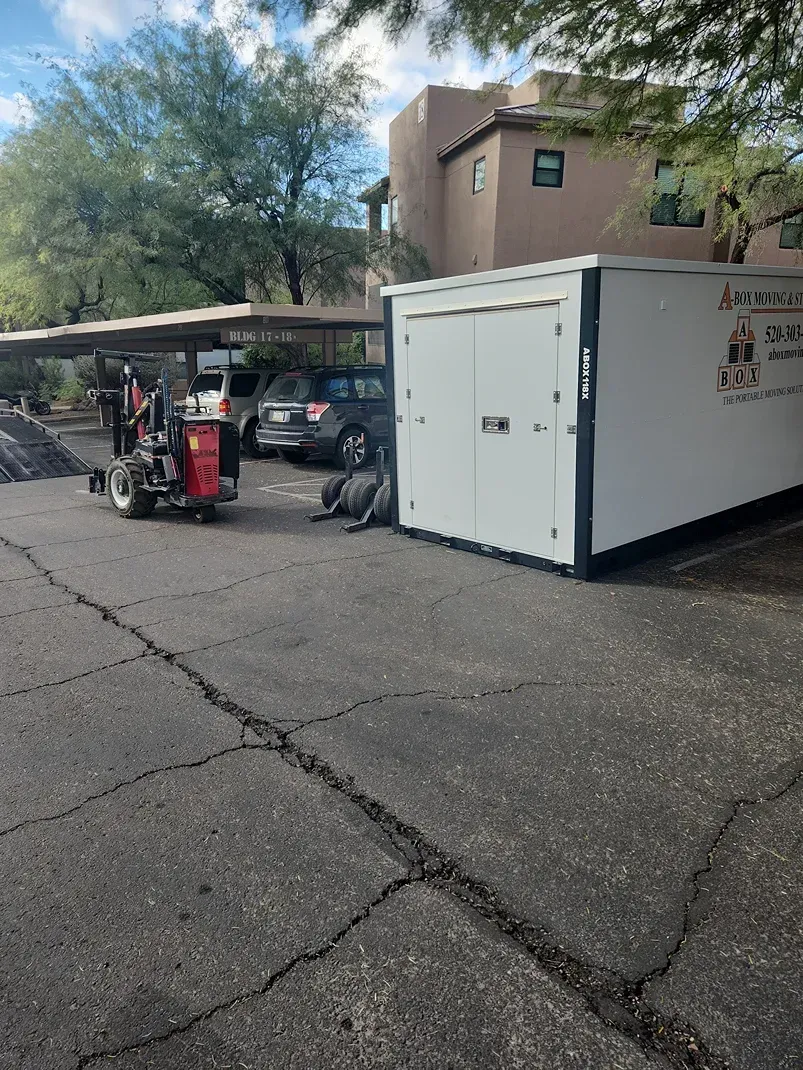 Forklift beside a white storage container in a parking lot near trees and a building.