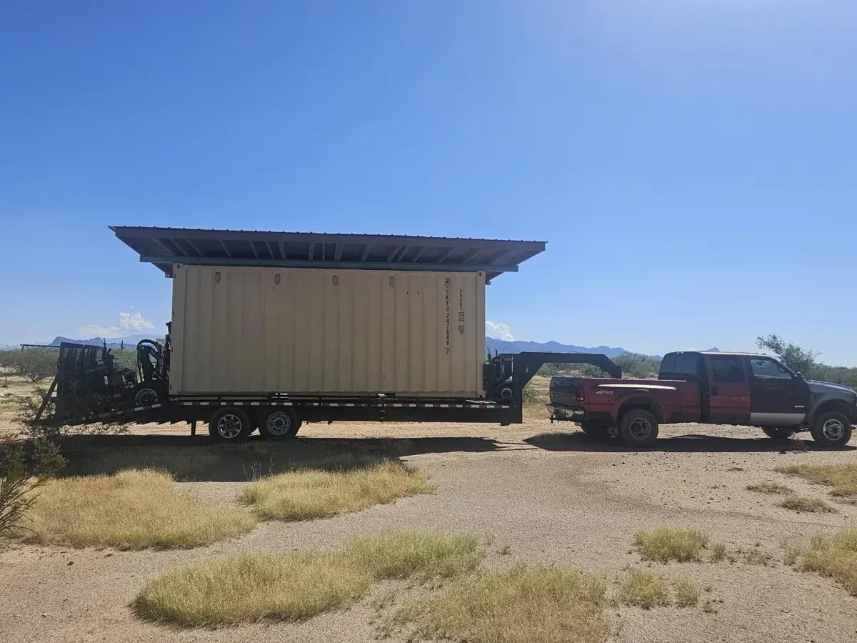 Red pickup truck towing a beige tiny house trailer in a dry, open field under a blue sky