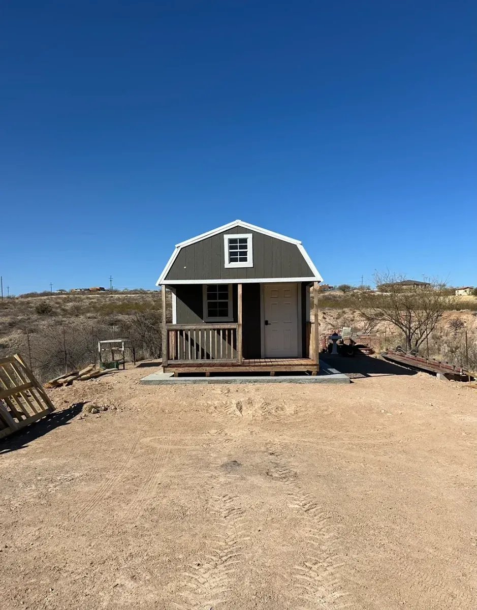 Small gray shed with white trim on a sandy lot under a clear blue sky.