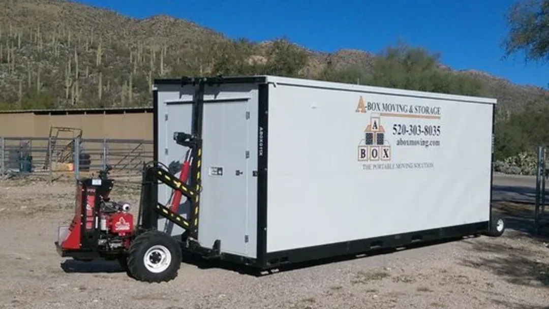 Red trailer-mounted portable building with logo, parked on dirt near a mountain road.