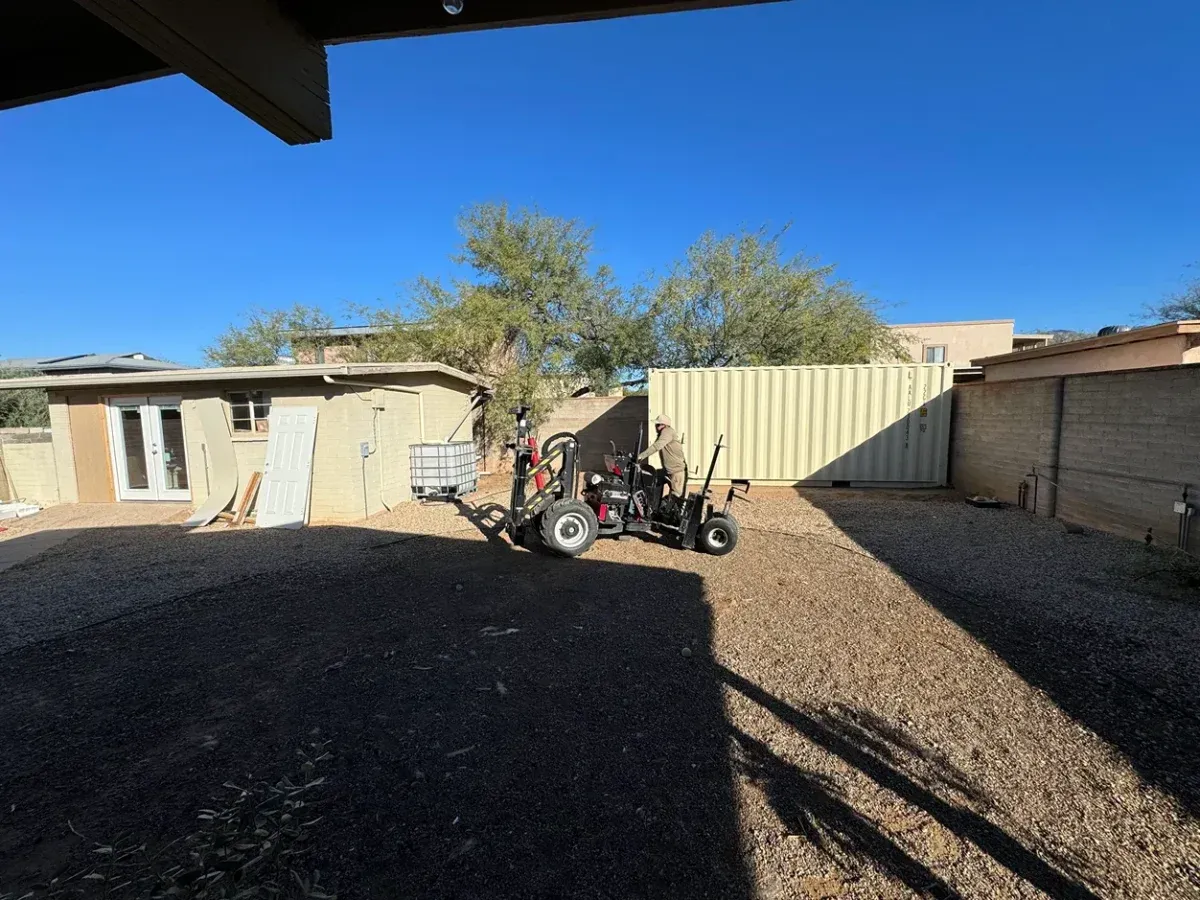 Sunlit gravel yard with a small motorcycle parked near a beige metal fence and house walls.