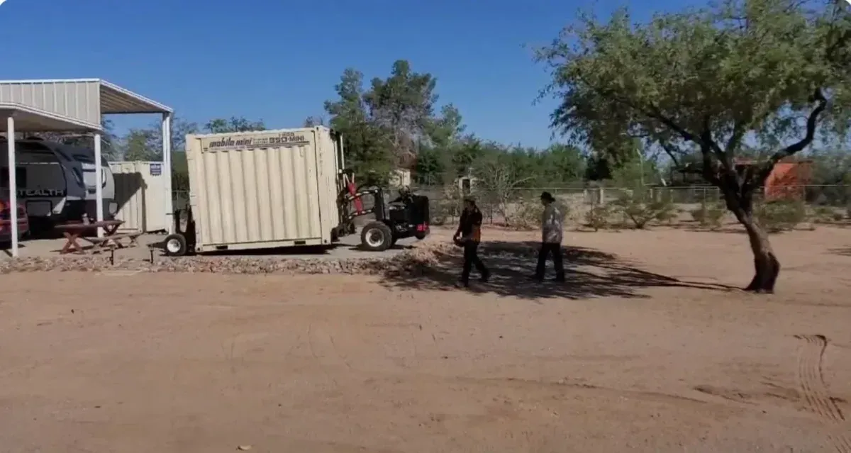 Two people walk across a dusty yard beside a beige container and a metal shed under a tree.