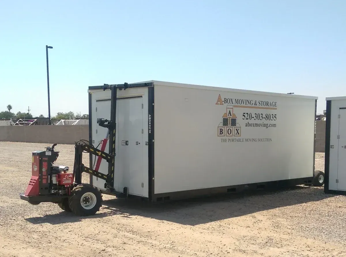 White storage trailer in a sandy lot, with a red bike-like machine beside it under a clear sky.